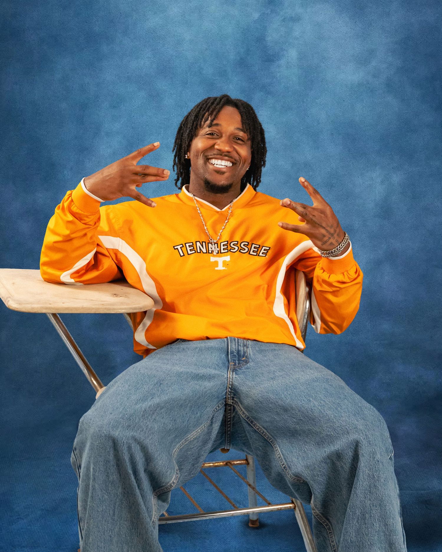 Smiling man in an orange Tennessee pullover and baggy jeans sits on a folding chair, flashing hand signs against a blue studio backdrop.