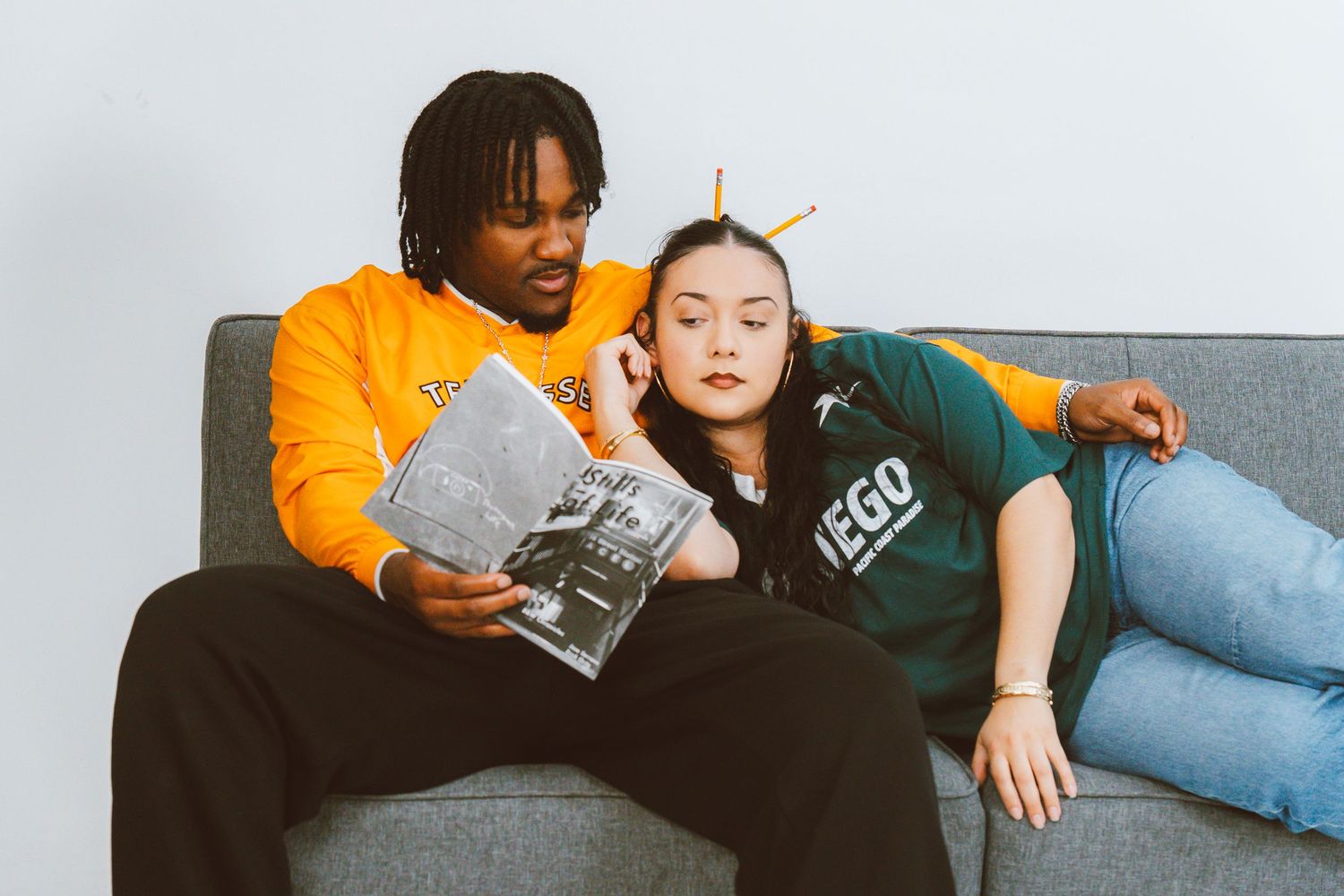 Young couple relaxing on a gray sofa as a man in an orange sweatshirt reads a magazine while a woman in a green tee rests against him.