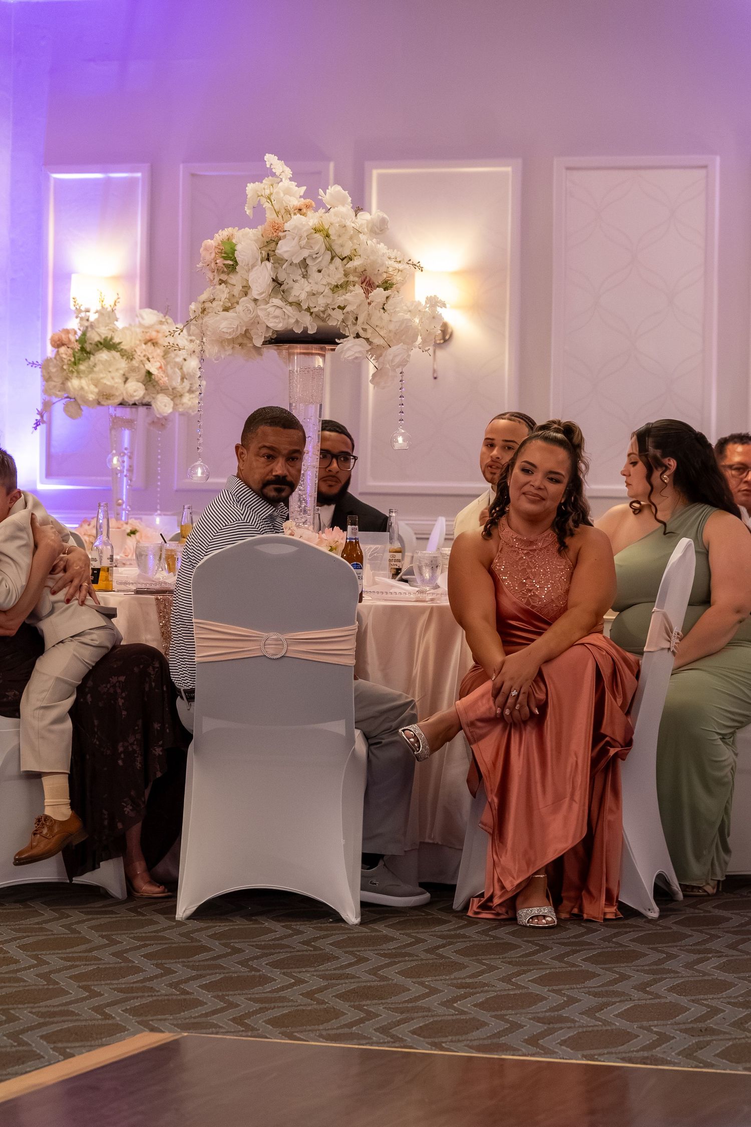 Elegantly dressed guests seated at a formal reception with tall white floral centerpieces and purple ambient lighting. Sweet 16/Quinceañera