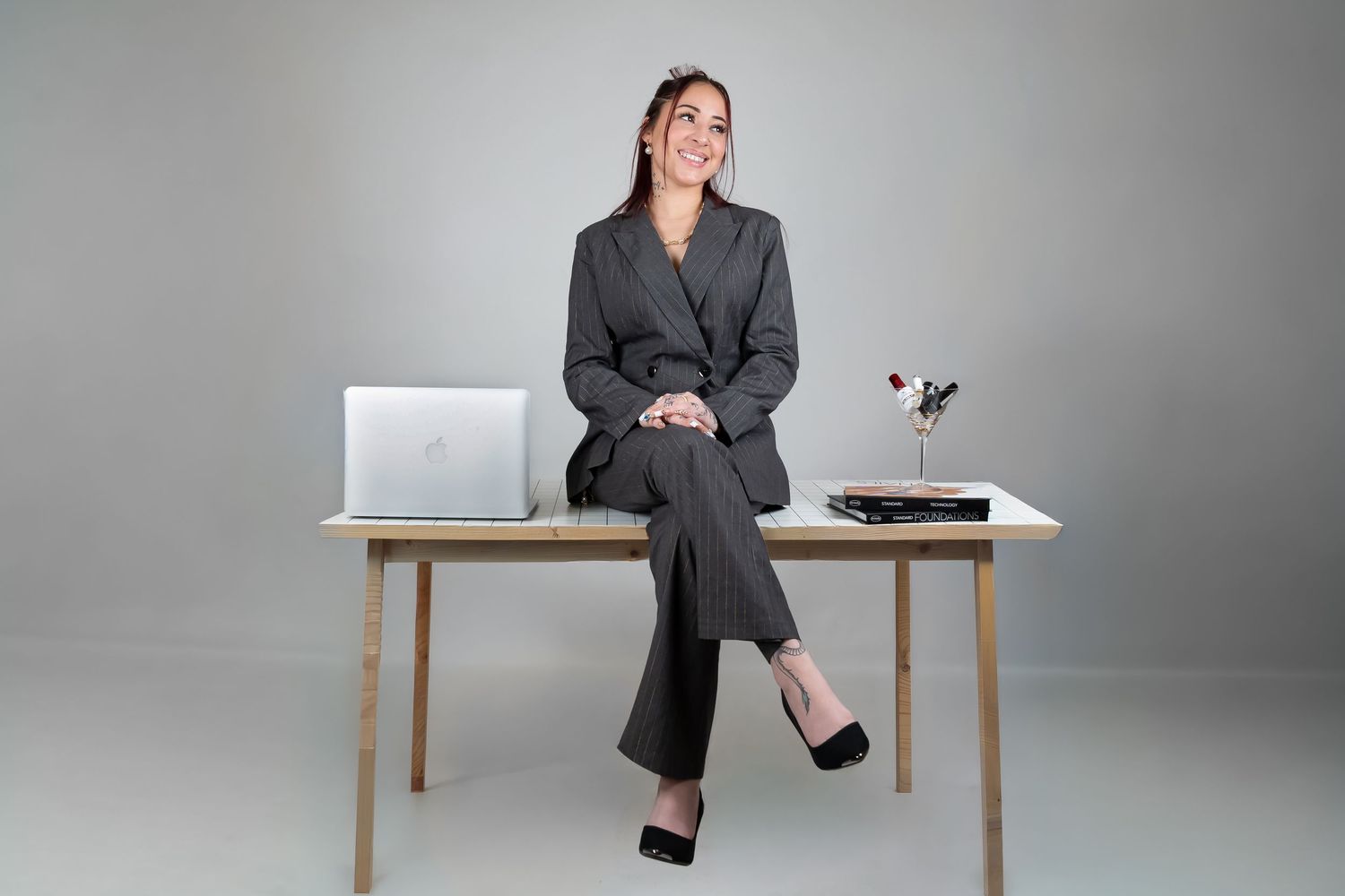 Confident businesswoman in gray suit sits on wooden desk with laptop in bright studio setting.