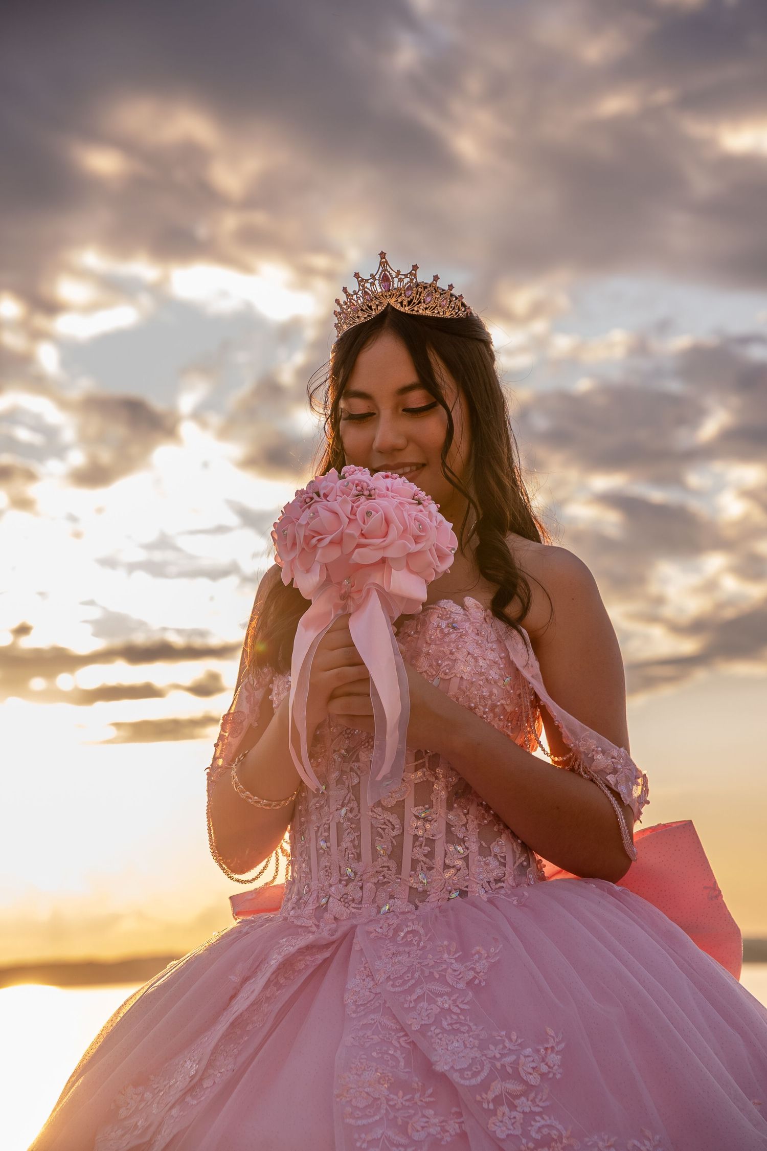 Quinceañera in pink lace gown and tiara smelling pink rose bouquet at golden sunset.