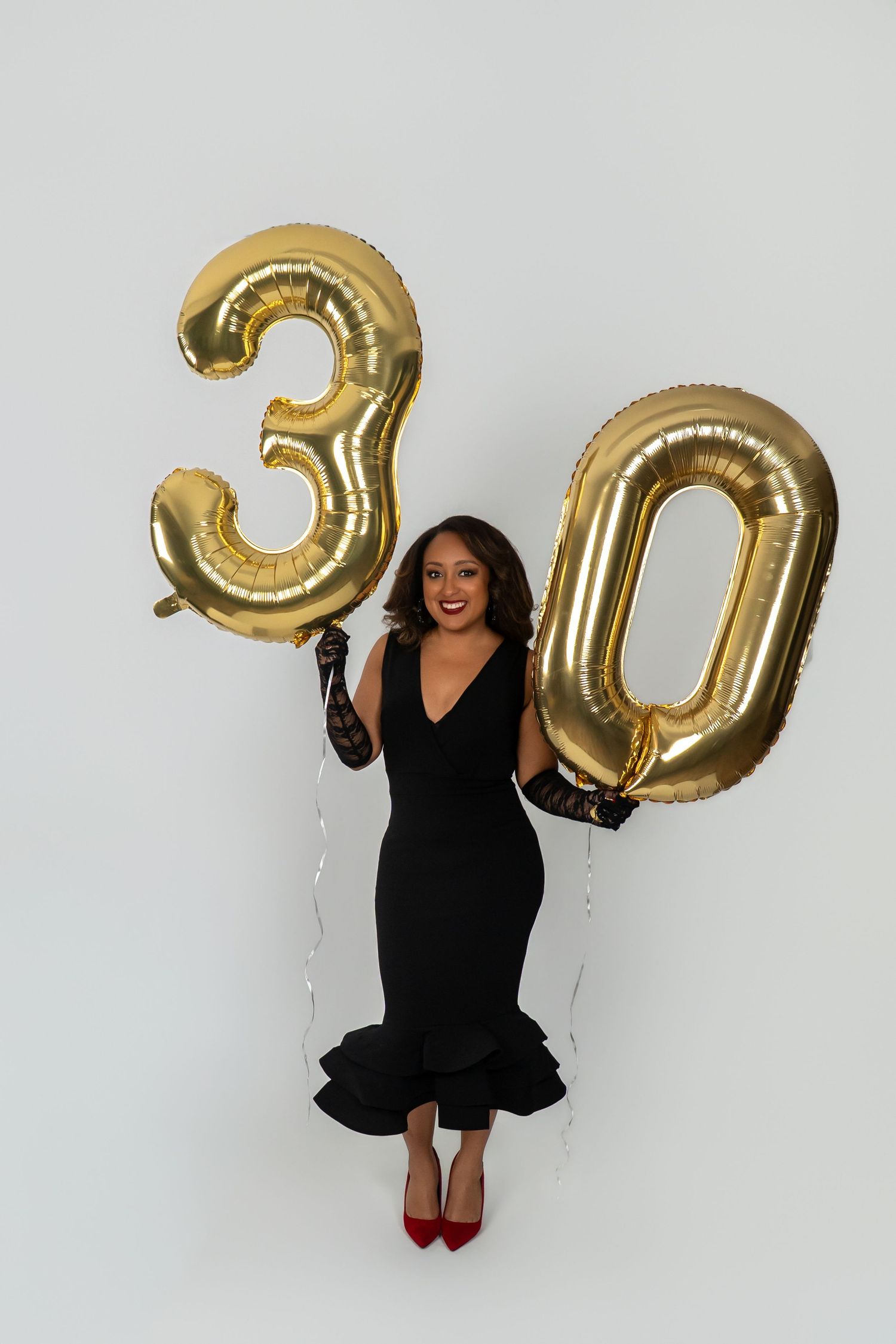 Woman in black dress holding gold number 30 balloons, celebrating her 30th birthday against white background.