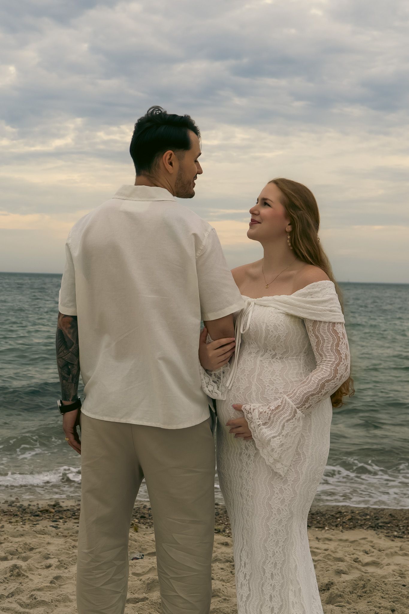 Pregnant couple in white and beige outfits sharing a romantic moment on a sandy beach at dusk.