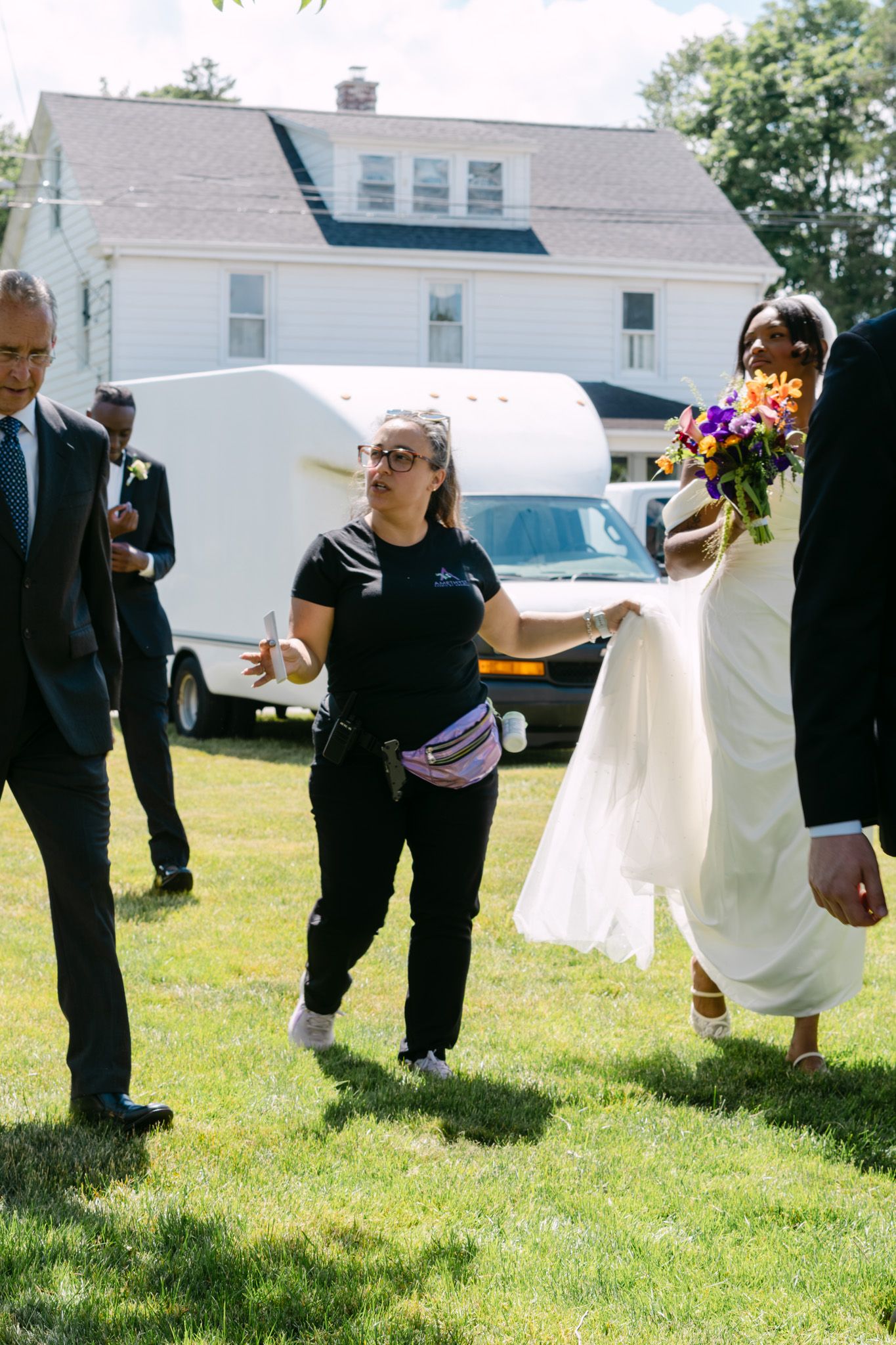 Wedding coordinator assists bride in white gown holding colorful bouquet outdoors on sunny day near white house.