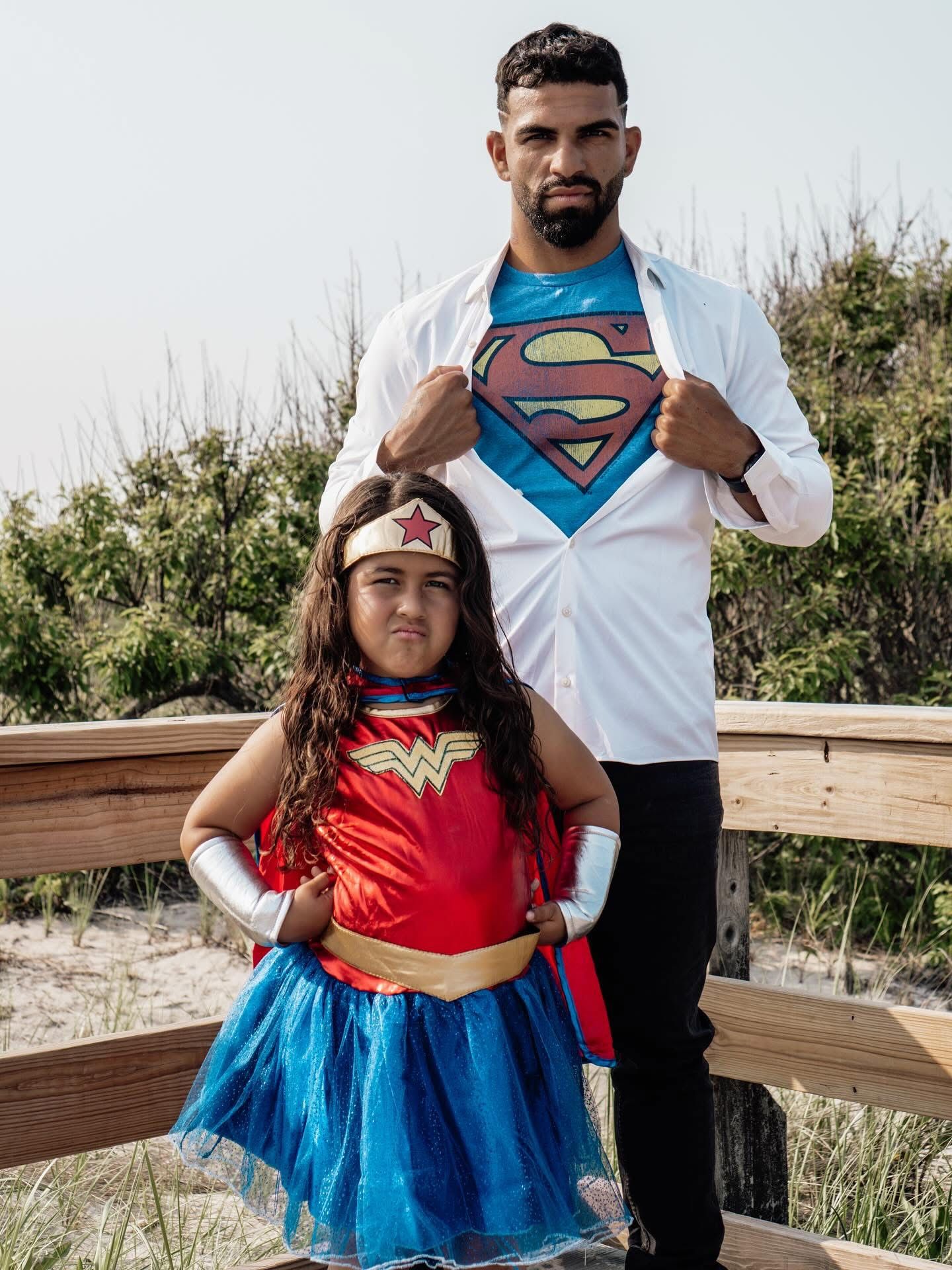 Man in Superman shirt poses with young girl in Wonder Woman costume on a wooden bridge outdoors.