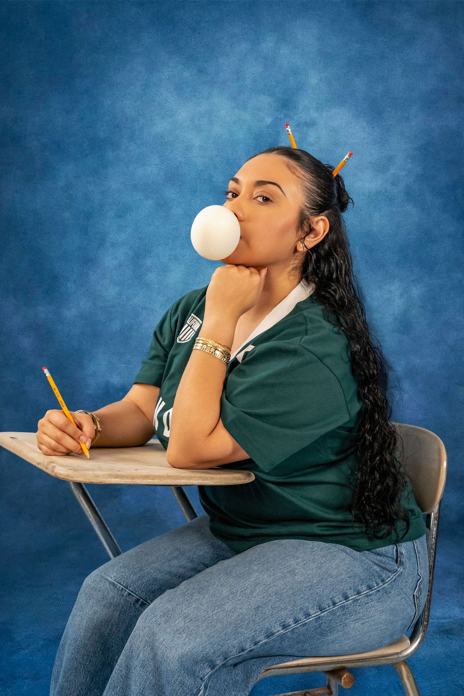 Woman in a green jersey and jeans blows a large white bubblegum bubble while sitting at a school desk with pencils in her hair.