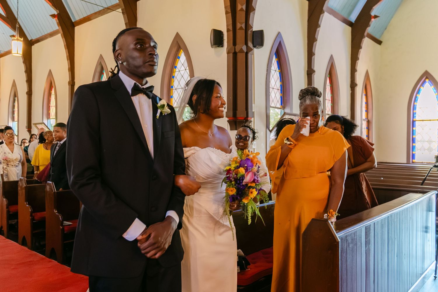 Bride and groom stand at the altar in a sunlit church during their wedding ceremony, surrounded by guests in colorful attire.