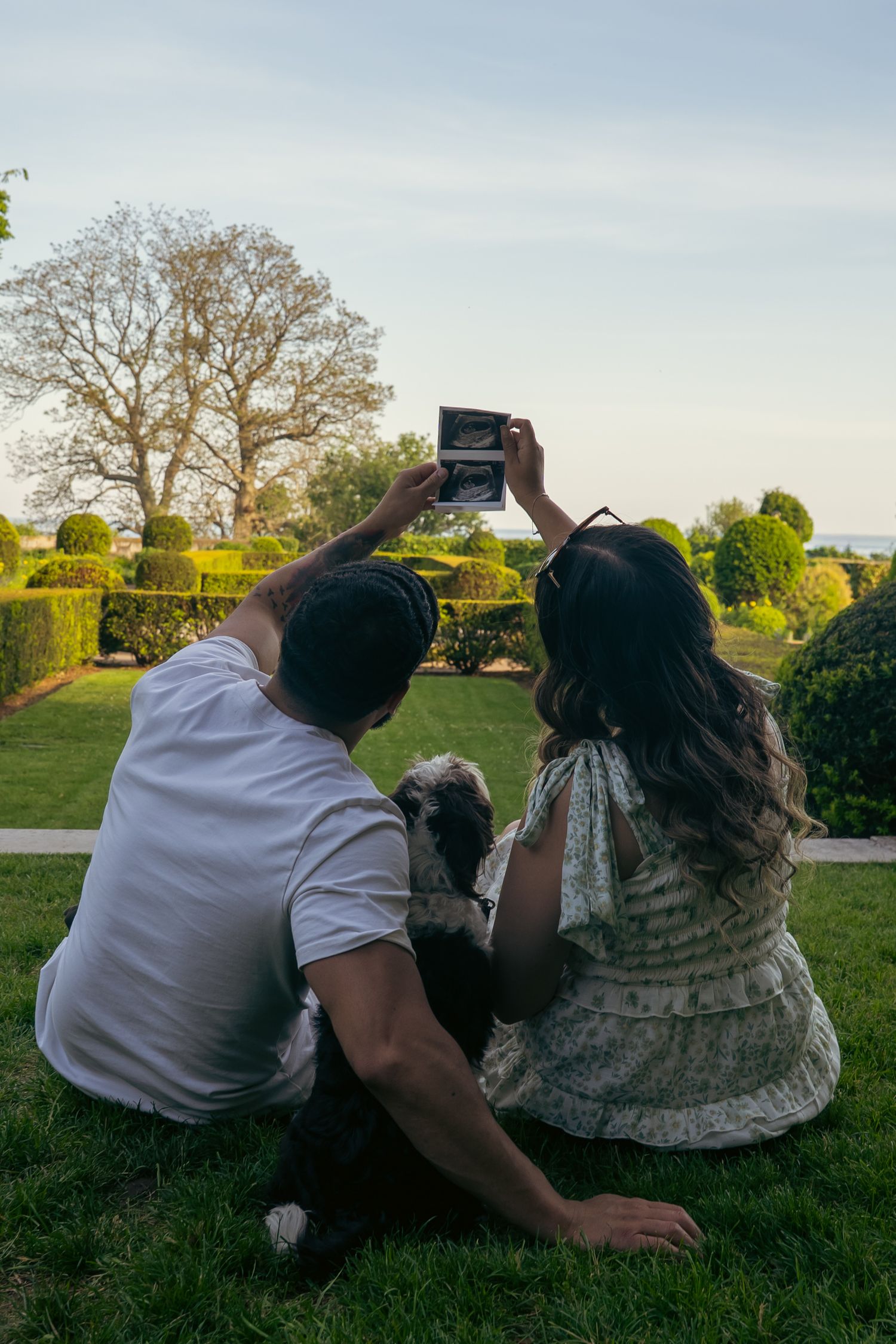 Couple sitting on grass with dog, holding ultrasound photo in a manicured garden at dusk.