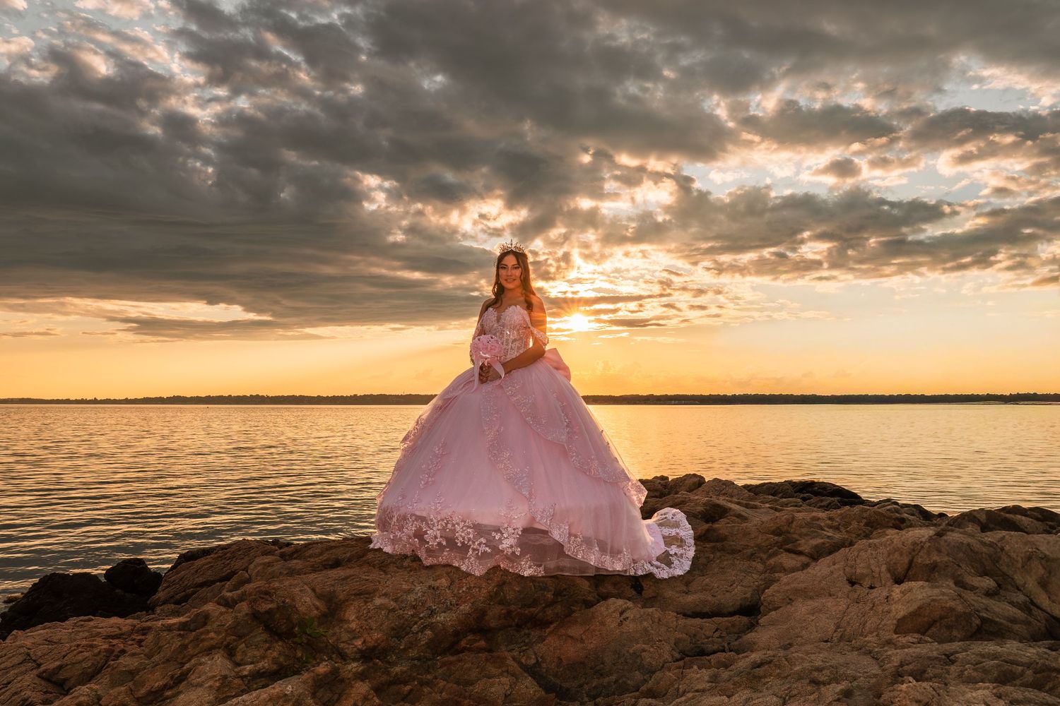 Girl in pink quinceañera gown poses on rocky shoreline at golden sunset over calm water.