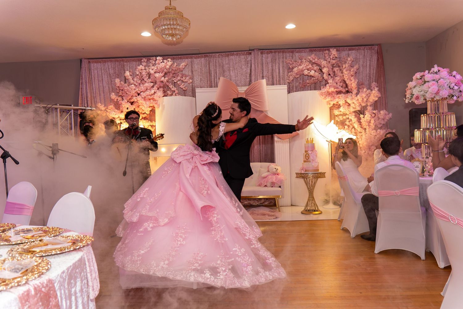 Quinceañera dancer in pink gown performs waltz amid dry ice fog before floral cherry blossom backdrop.