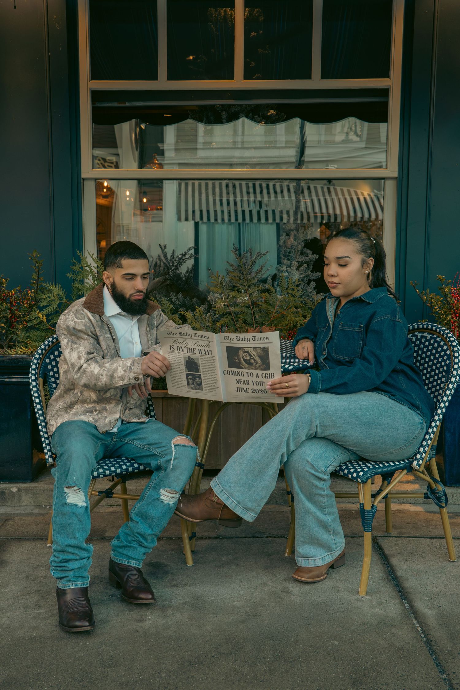 Couple reads newspaper together at outdoor cafe table with blue storefront background.