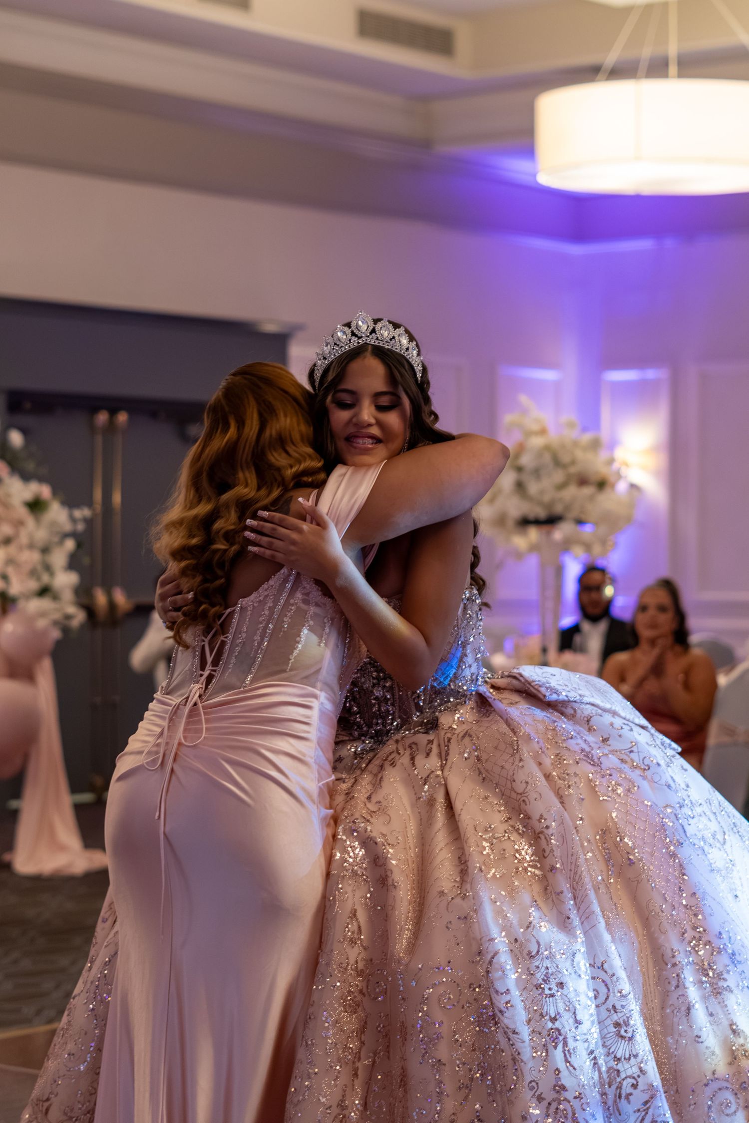 Two women in elegant gowns share a heartfelt hug at a quinceañera, one wearing a sparkling ball gown and tiara. Sweet 16/Quinceañera