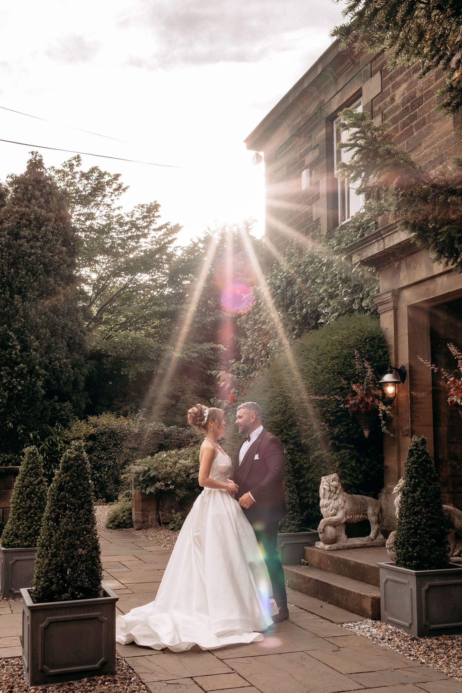 Bride and groom stand together in golden sunlight outside a stone manor, surrounded by lush gardens and topiary.