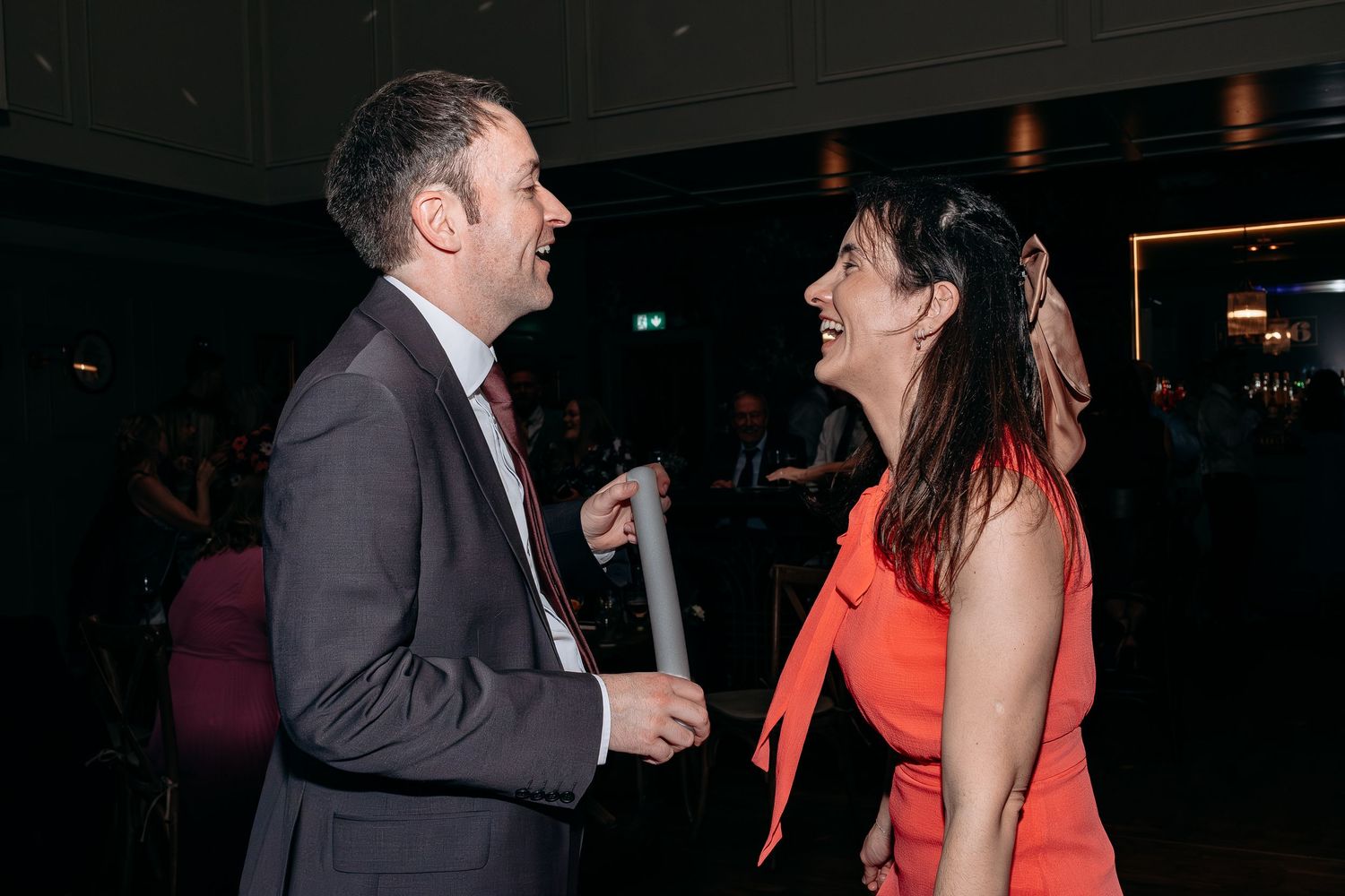 A man in a suit and woman in a coral dress laugh together at a formal evening social event.