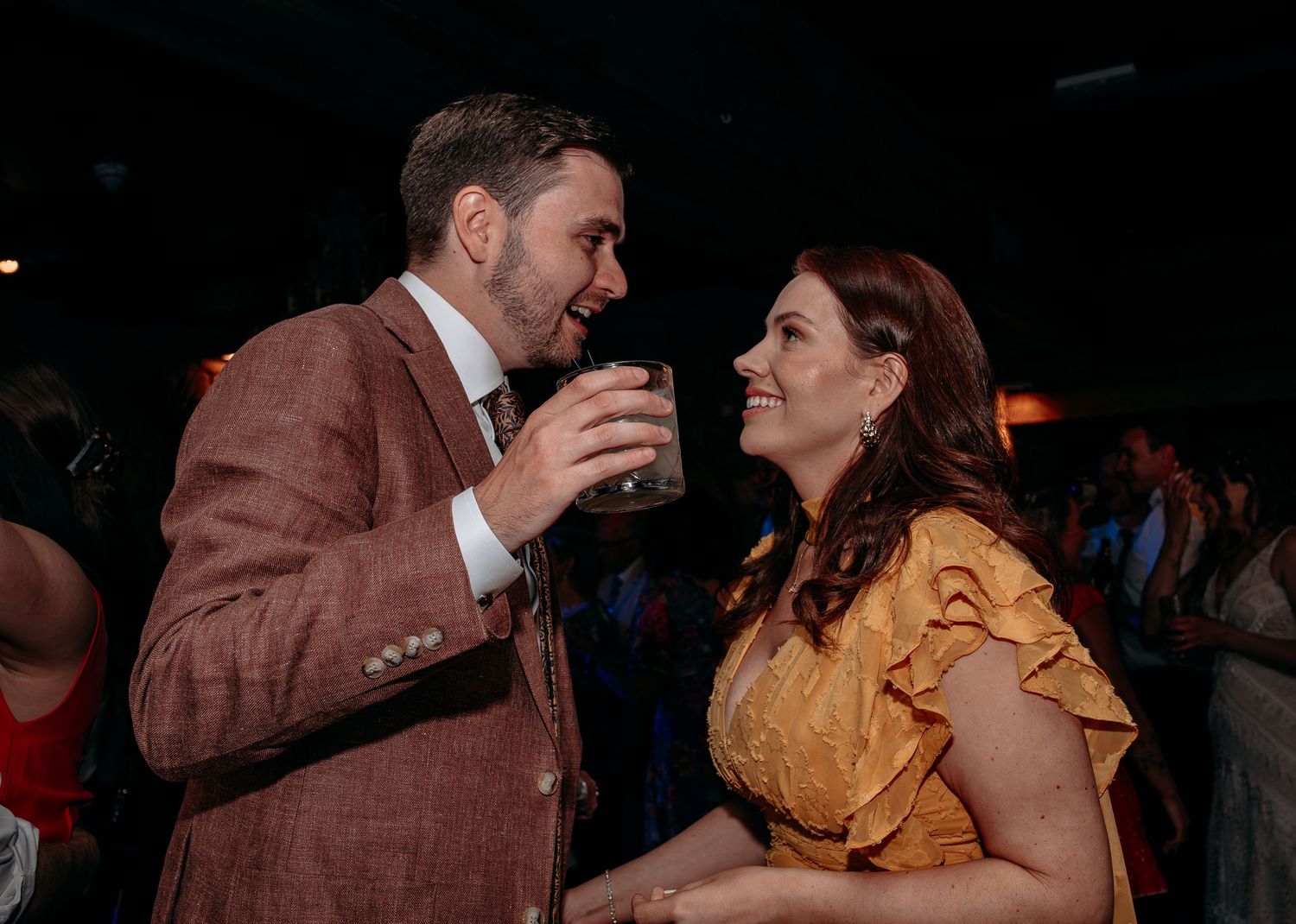 A man in a brown suit and woman in yellow dress share a romantic moment at a party event.