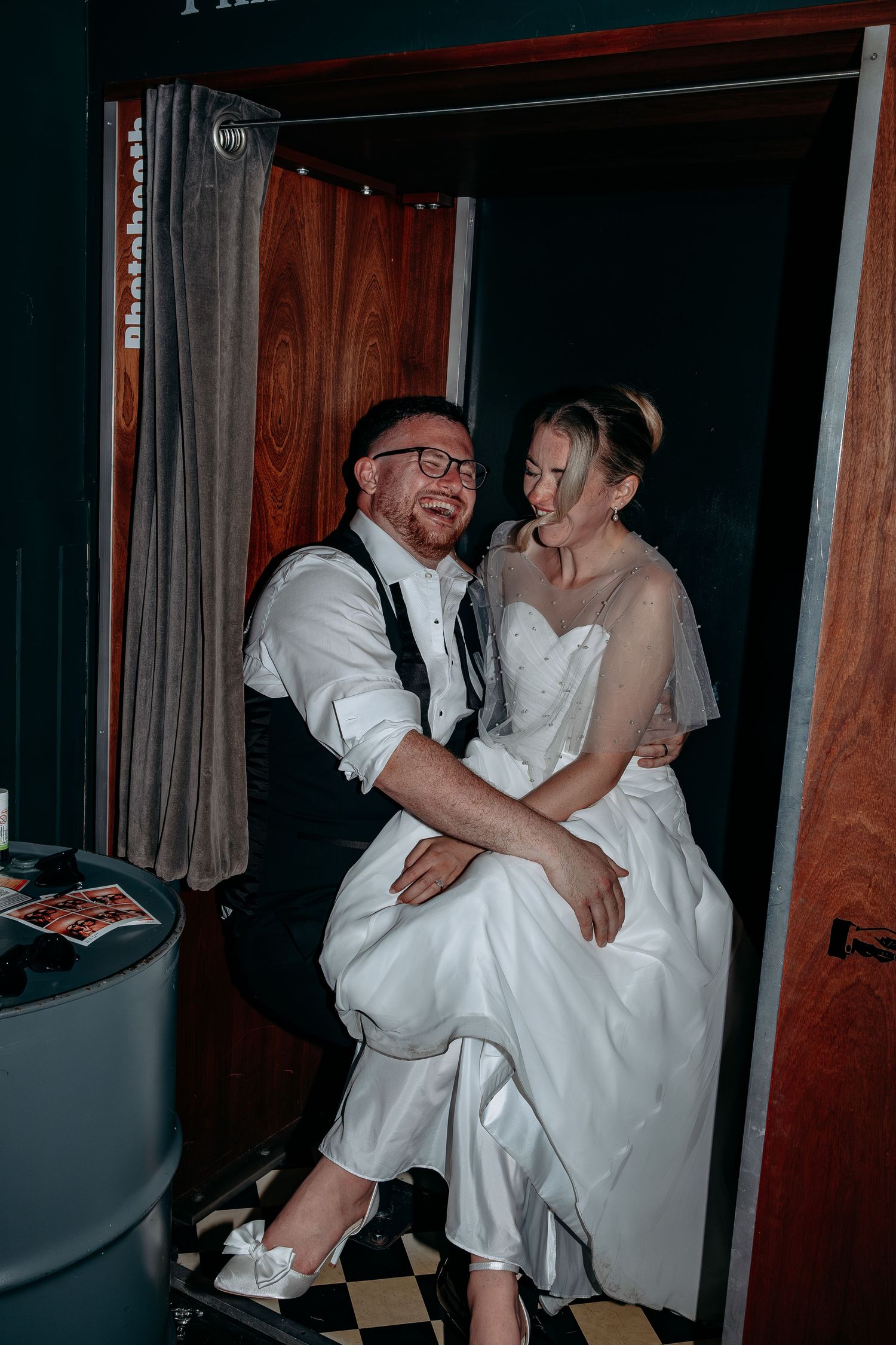 Happy newlyweds laughing together in a photo booth, bride in white gown sitting on groom's lap.