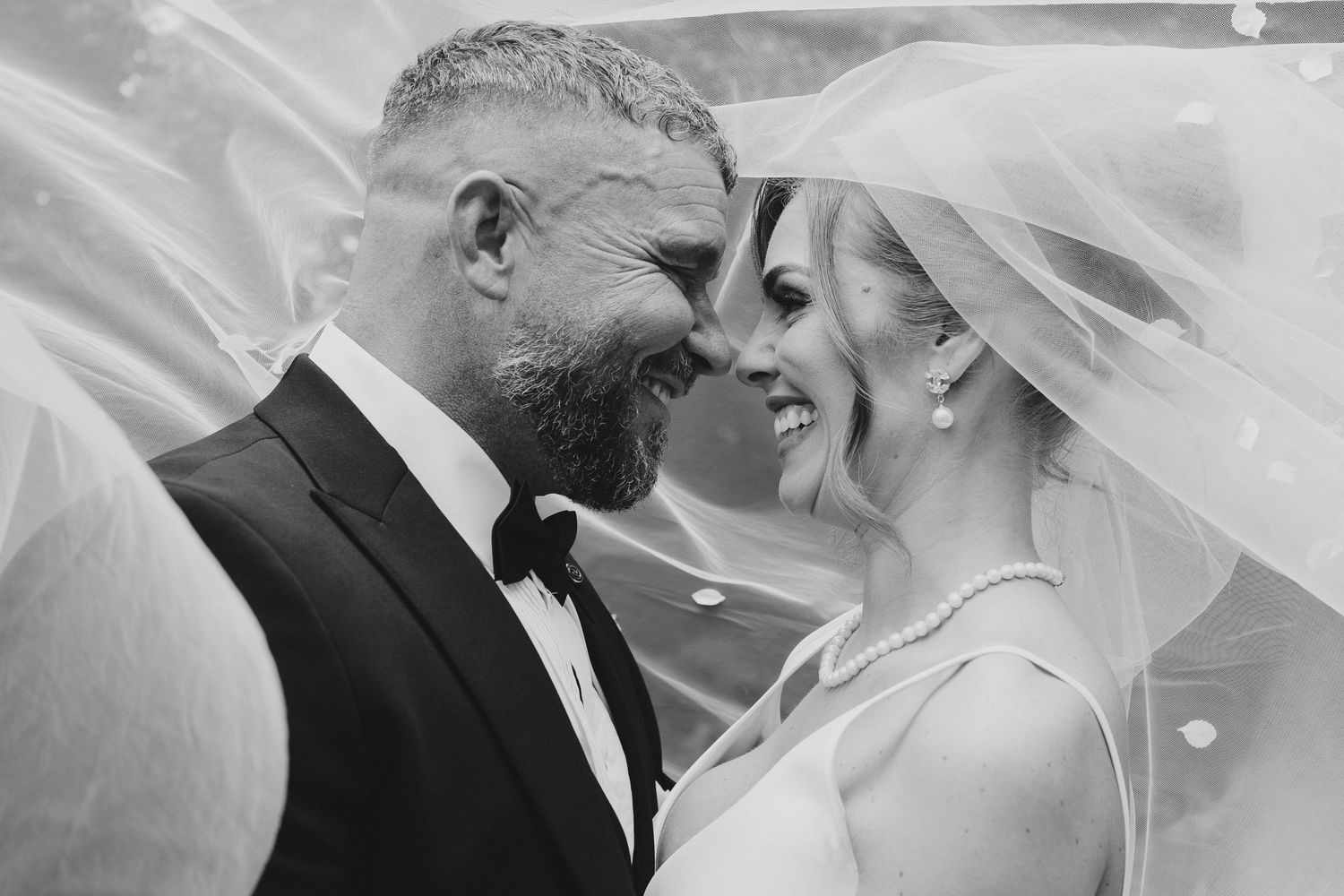 Black and white photo of smiling bride and groom touching foreheads under wedding veil.