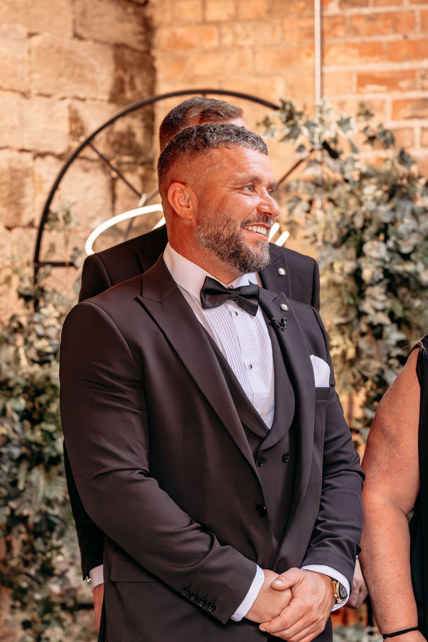 Smiling groom in black tuxedo with bow tie stands at wedding altar decorated with greenery and brick wall backdrop.