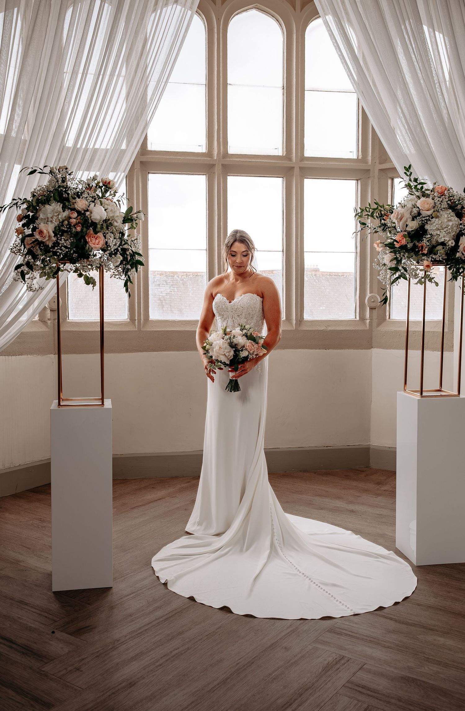 Bride in strapless white gown holding bouquet stands between floral pedestals in elegant venue.