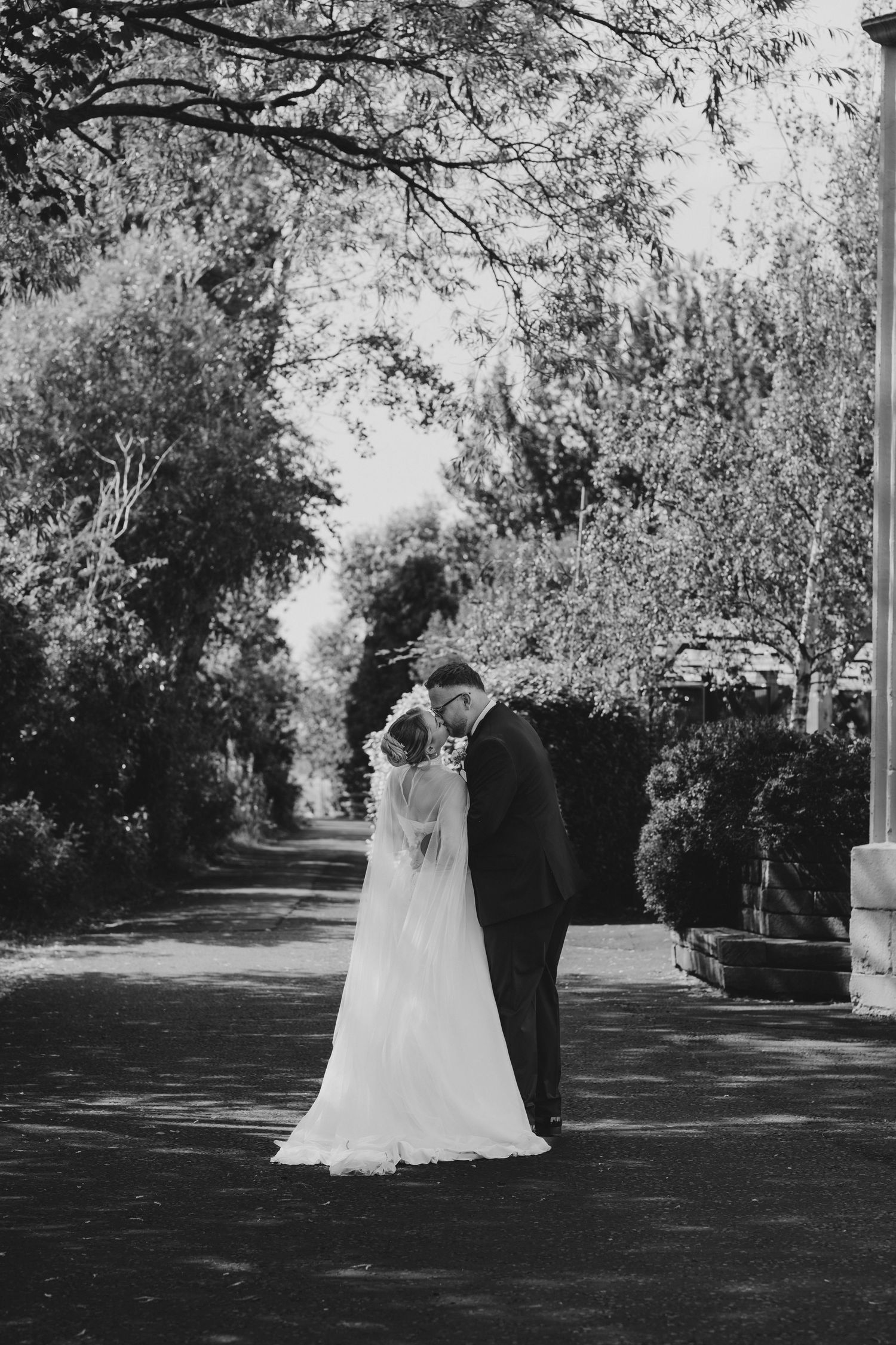 Black and white photo of a bride and groom sharing an intimate moment on a tree-lined path on their wedding day.