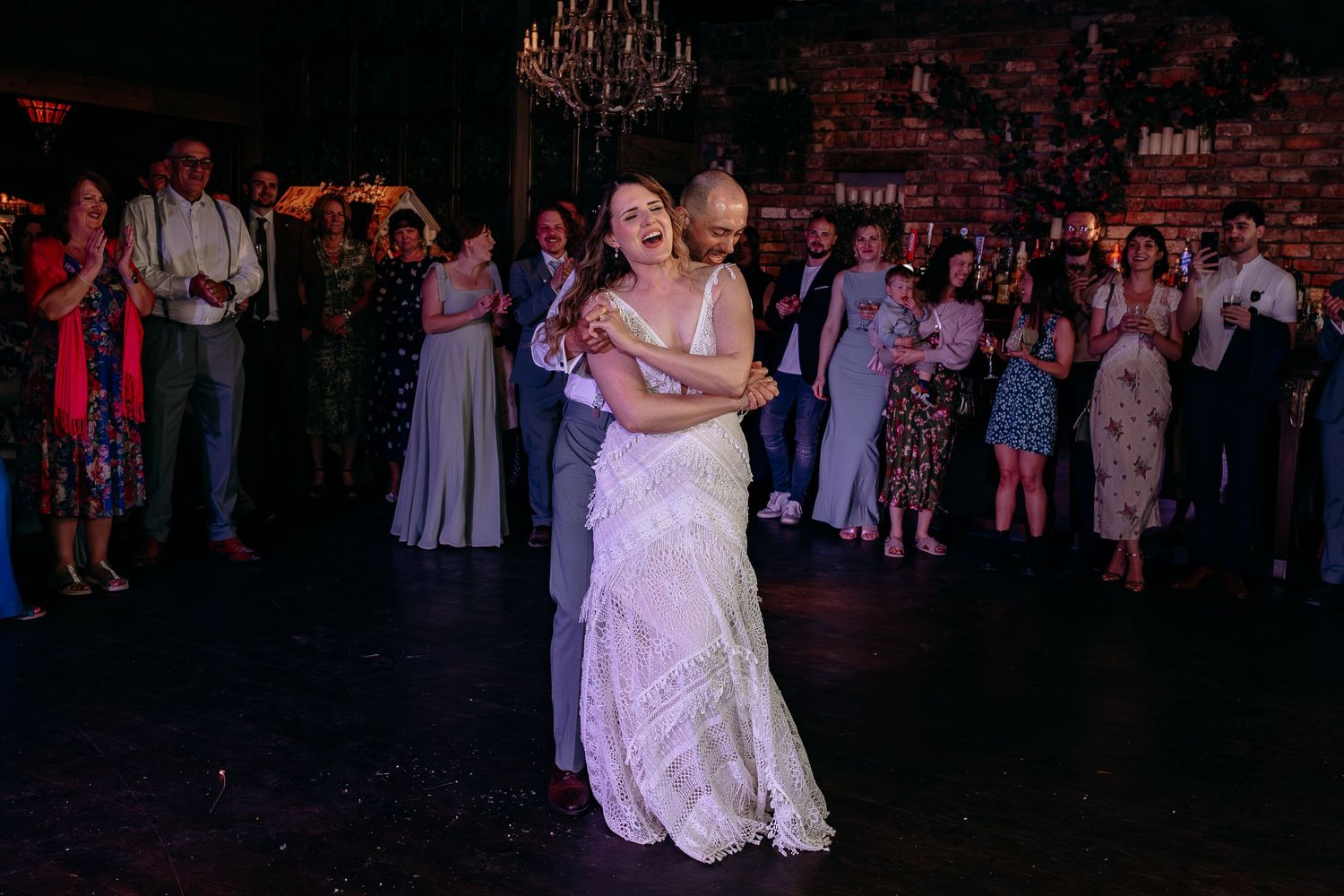 Bride and groom share their first dance surrounded by smiling wedding guests in a warmly lit rustic venue.