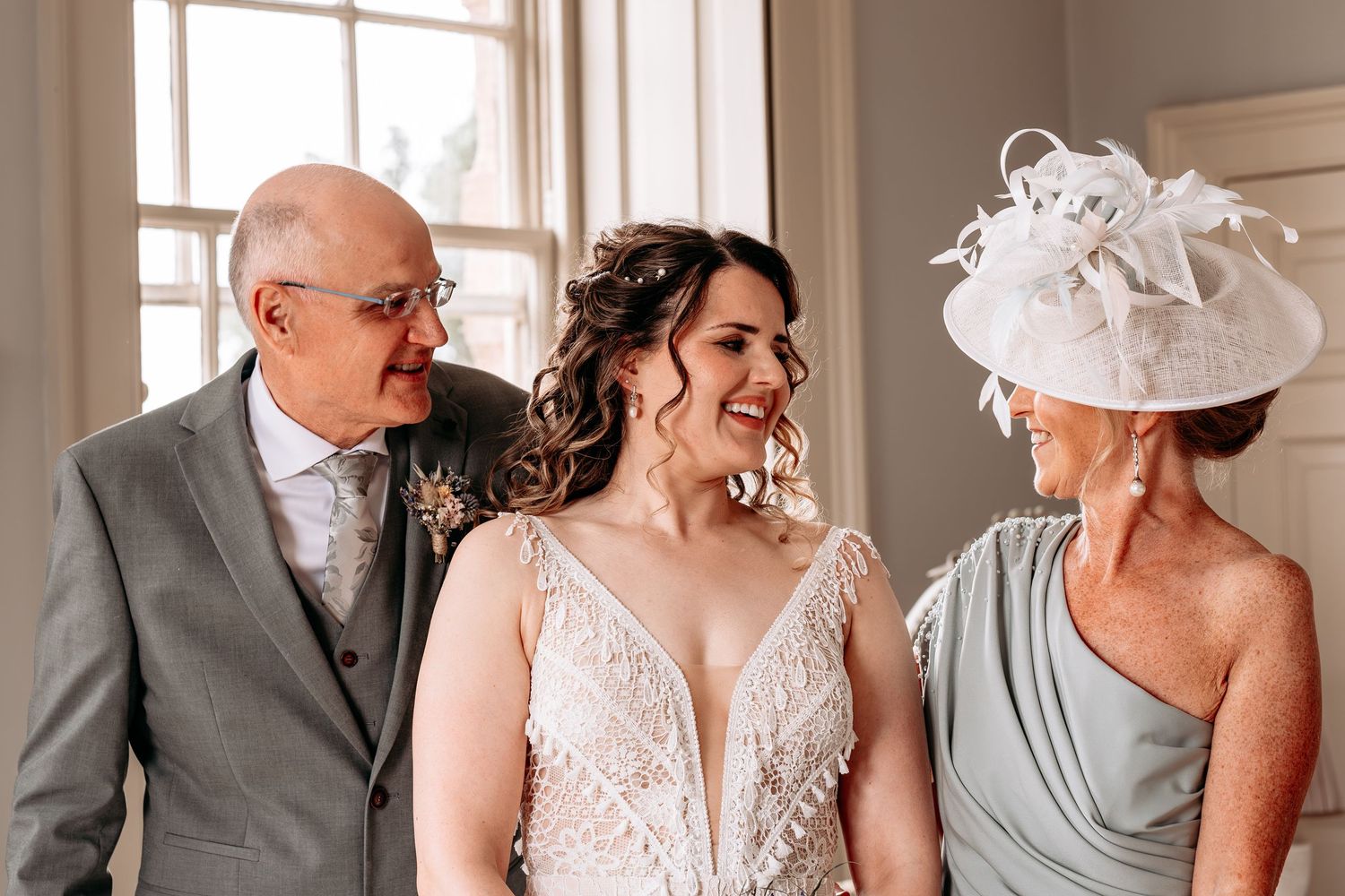 Bride in lace wedding dress smiles with elderly parents, mother wearing sage green gown and white fascinator hat.