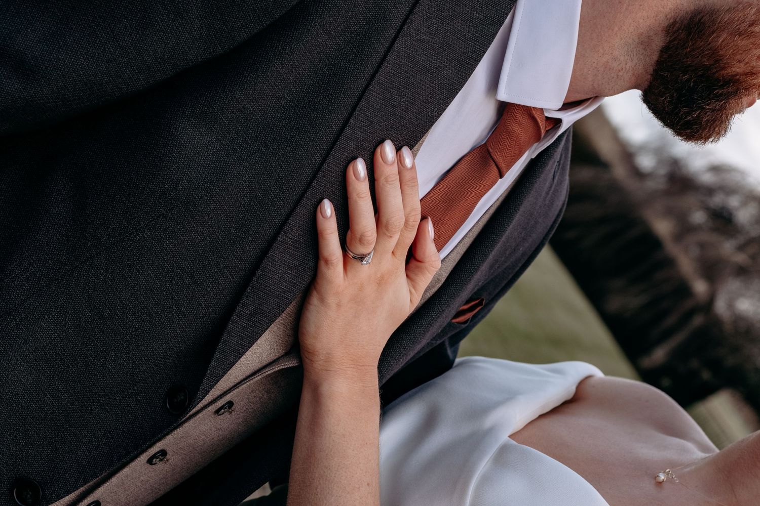Close-up of bride placing hand on groom's suit jacket, showing wedding ring detail at outdoor ceremony.