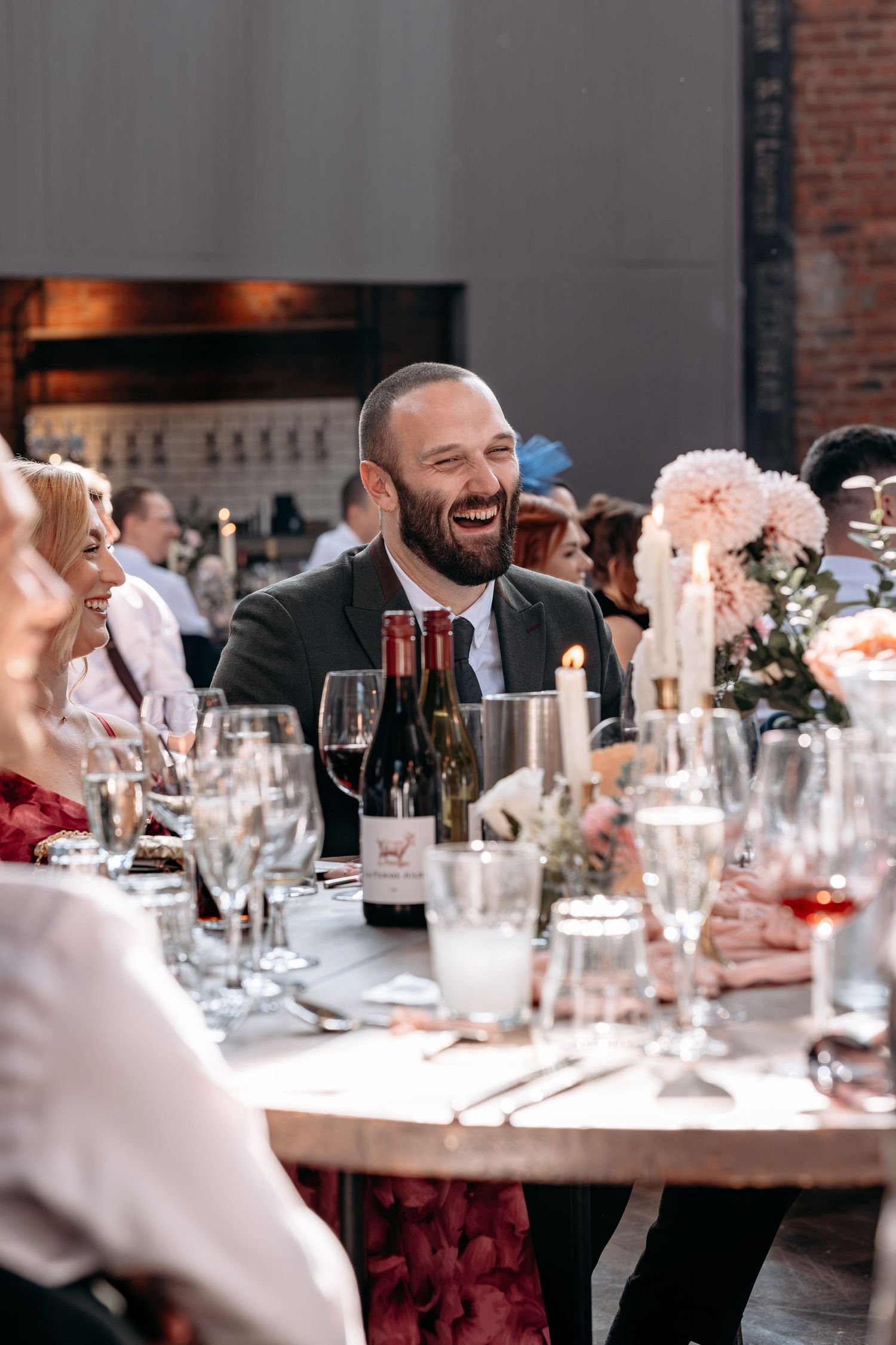 A laughing man in a dark suit sits at an elegantly set wedding reception table with wine bottles and floral centerpieces.