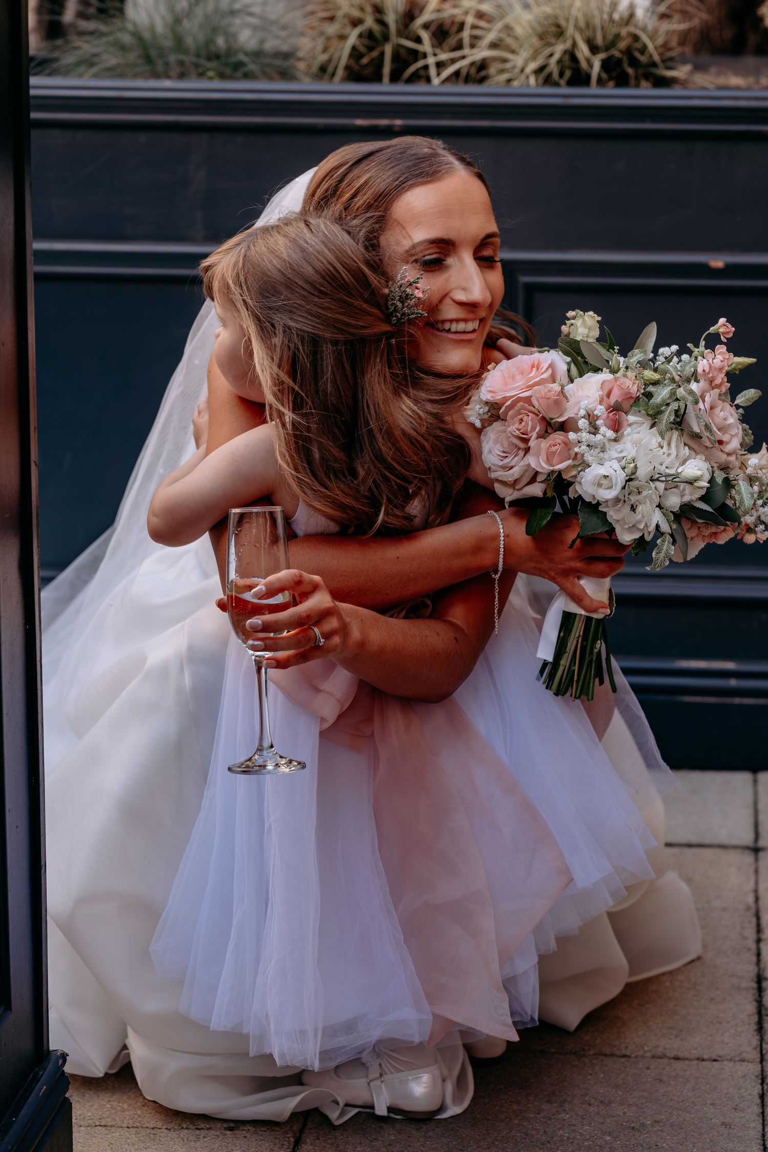 Bride in white gown hugs young flower girl while holding champagne flute and pink rose bouquet outdoors.