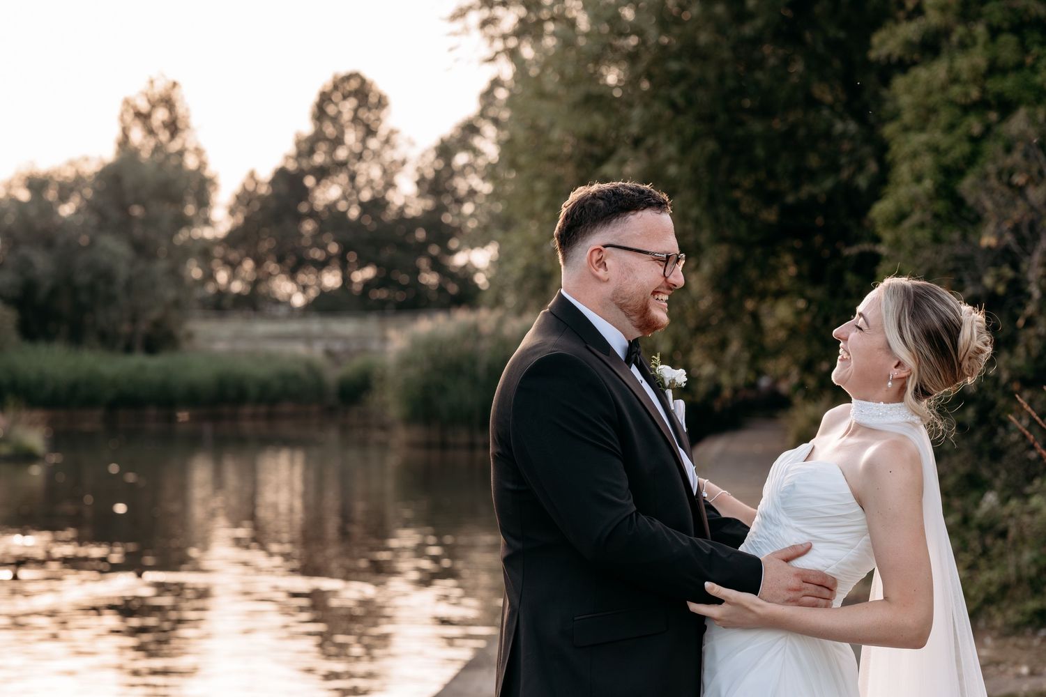 Bride and groom share a romantic moment by a serene lake at golden hour, surrounded by lush green trees.