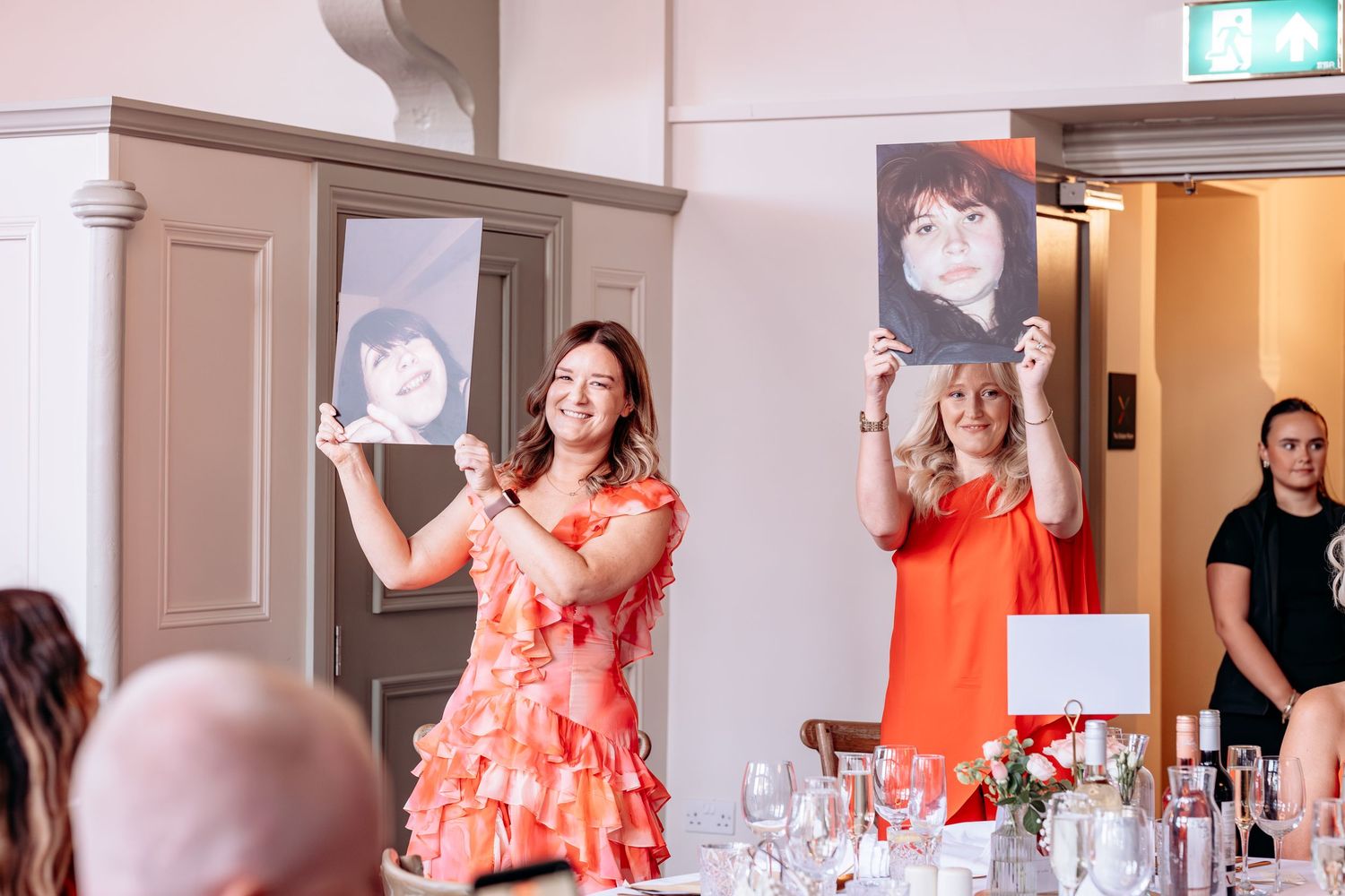 Two women in orange dresses hold up portrait photos at an elegant indoor event with decorated tables.