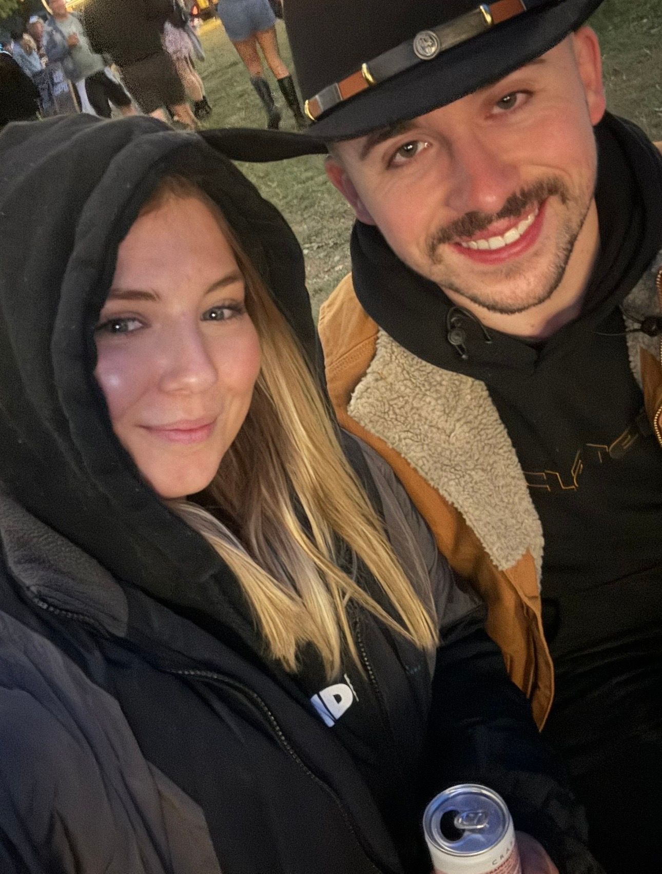 A smiling couple poses for a selfie at an outdoor festival, woman in black hoodie and man wearing a cowboy hat holding a can.