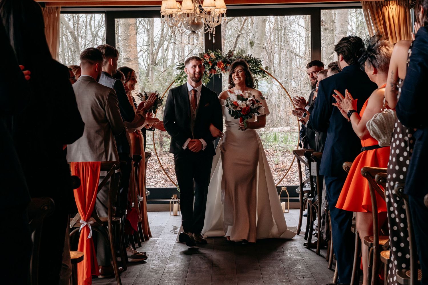 Bride and groom walk down the aisle together at an elegant indoor wedding ceremony with guests applauding.
