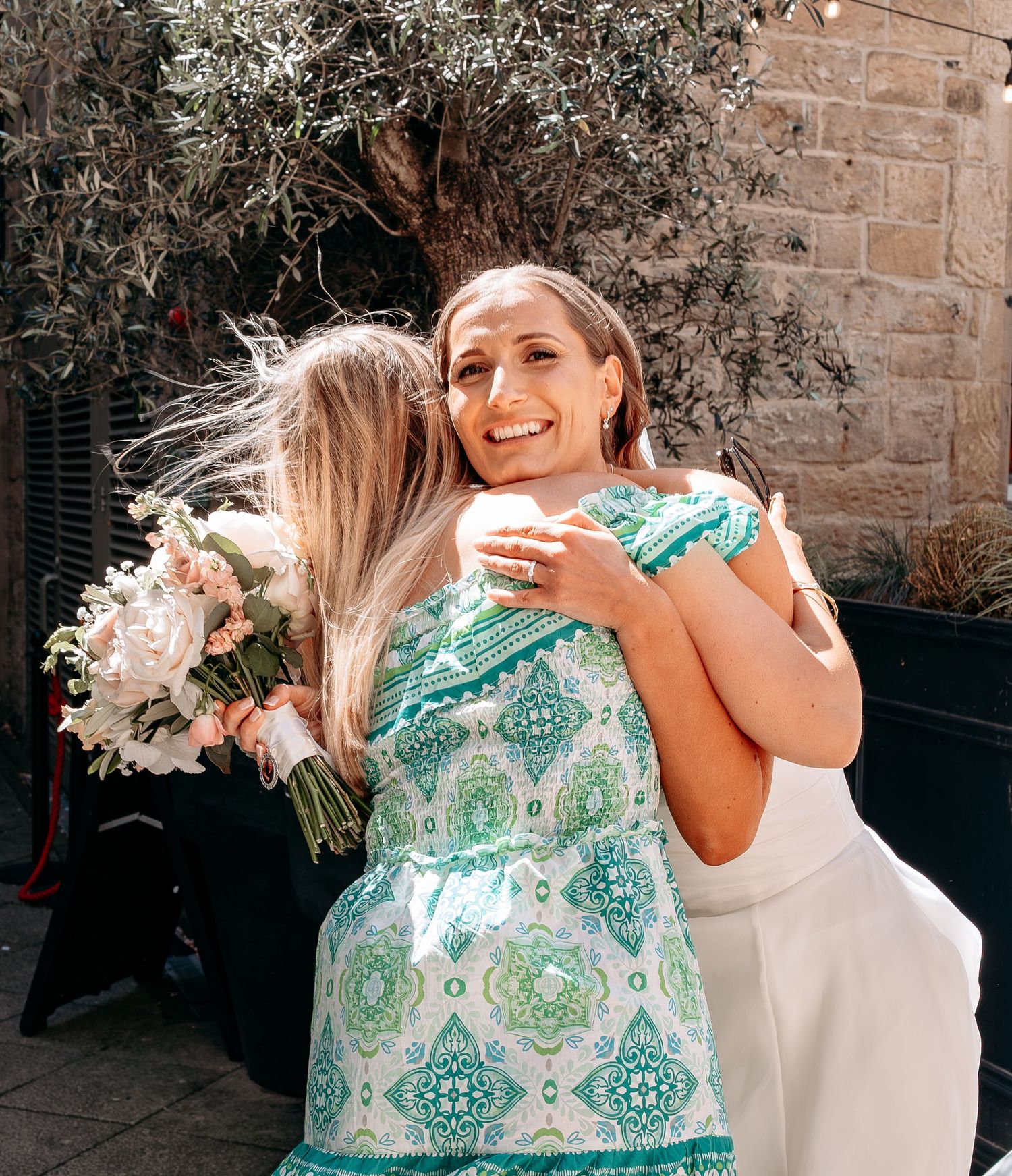 Two women embracing at a wedding, one in a green patterned dress holding a bouquet of white flowers outdoors.