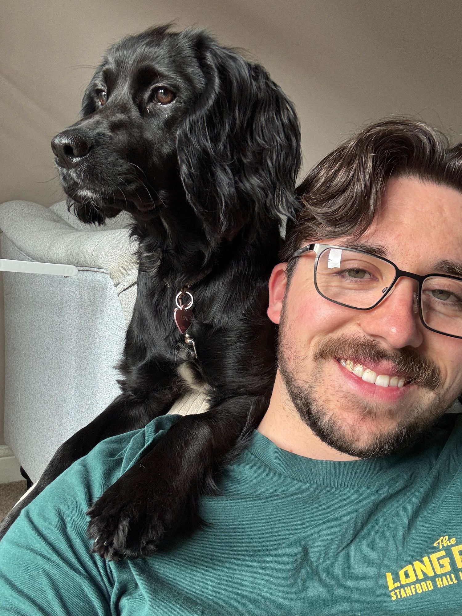 Man with glasses and beard smiling in selfie with black Cocker Spaniel dog wearing red collar indoors.