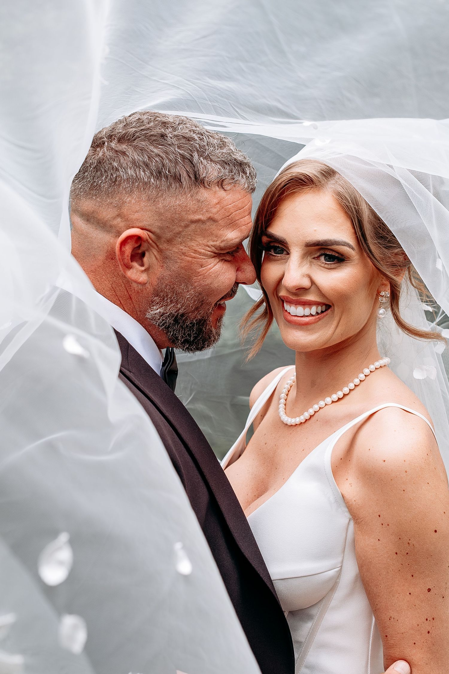 Bride and groom share a romantic moment under a flowing veil, bride smiling in white dress and pearl necklace.
