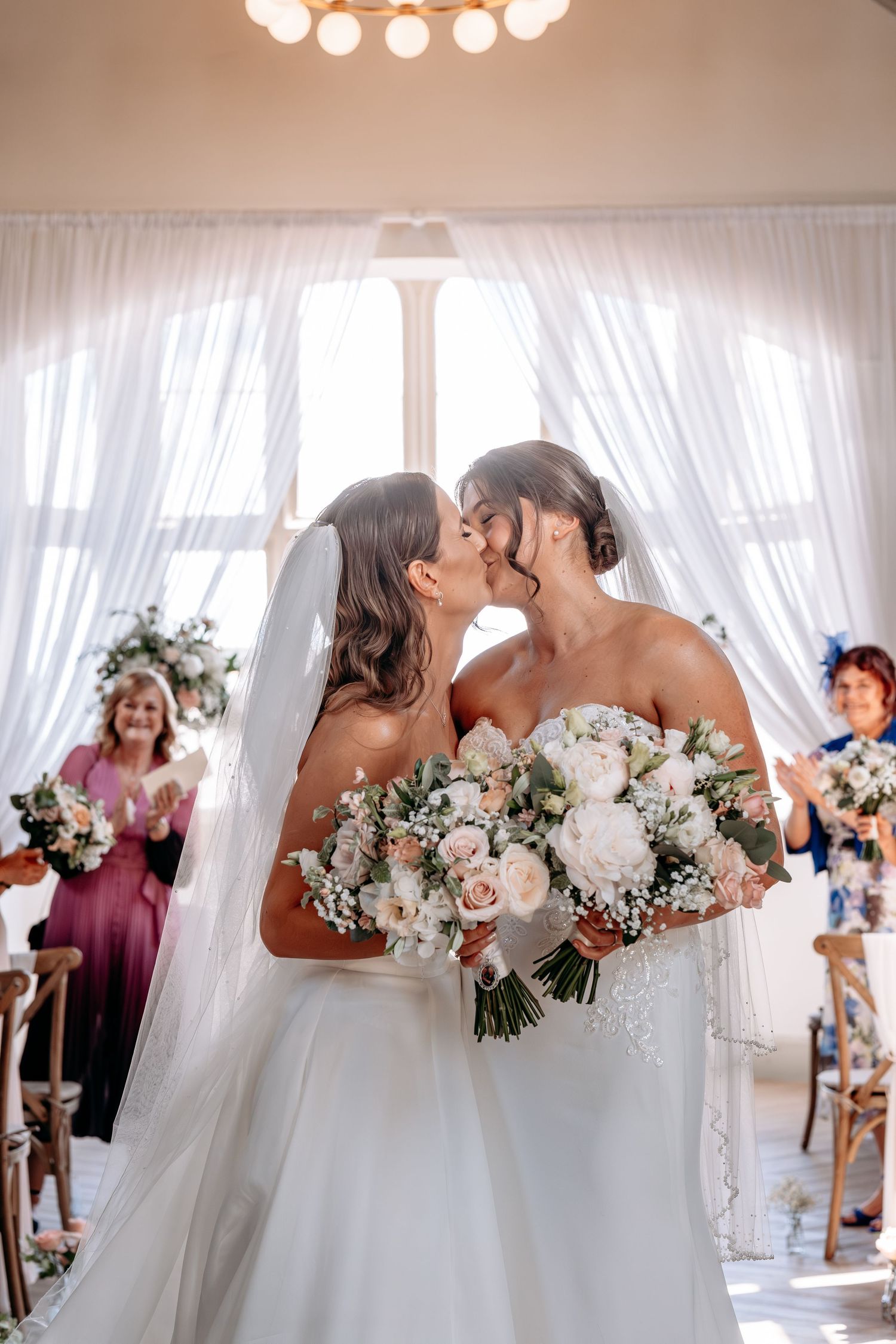 Two brides share a first kiss at their wedding ceremony, holding white and blush bouquets with guests applauding behind them.