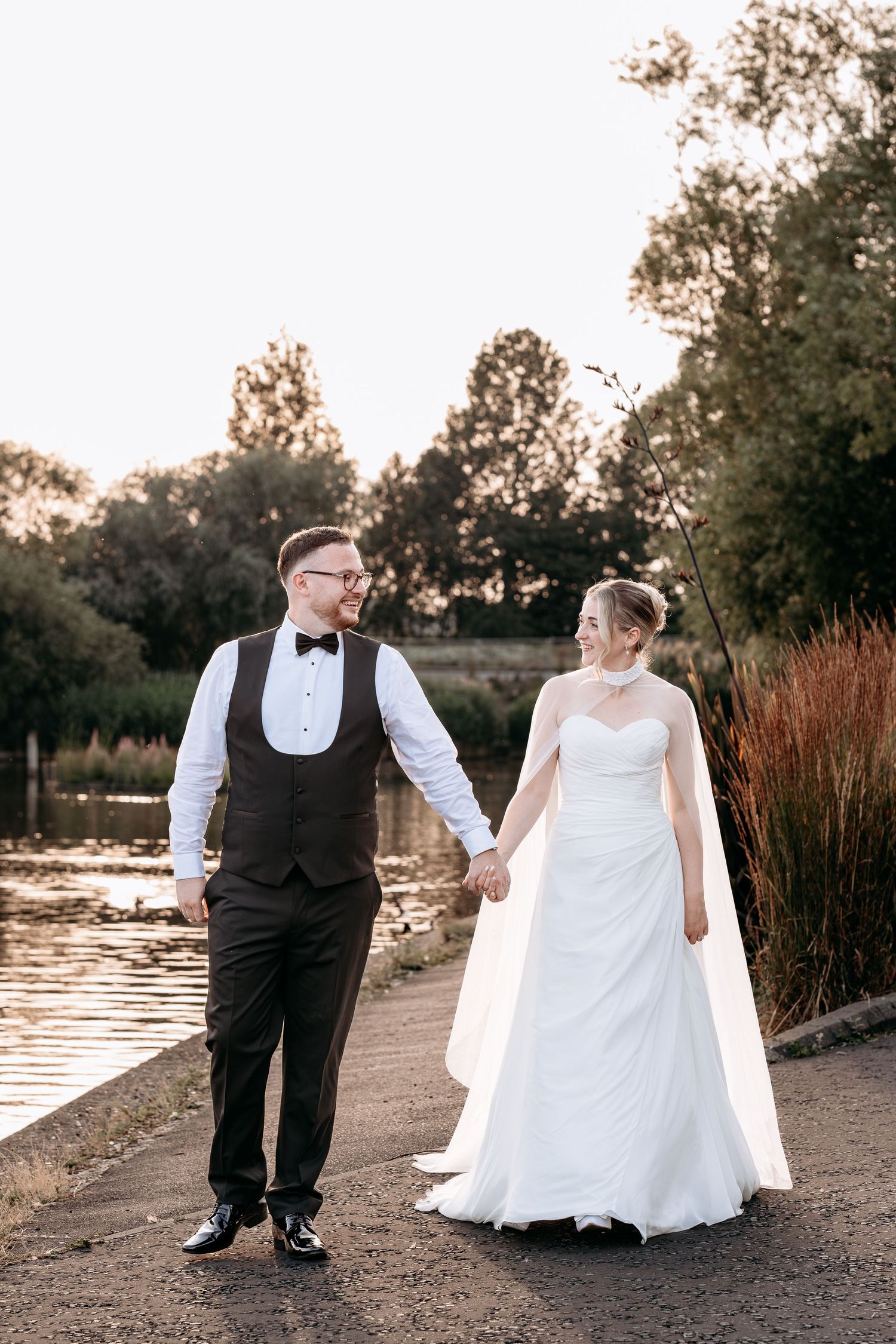 Bride and groom holding hands walking by a lakeside at sunset, groom in black tuxedo vest and bride in white strapless gown.