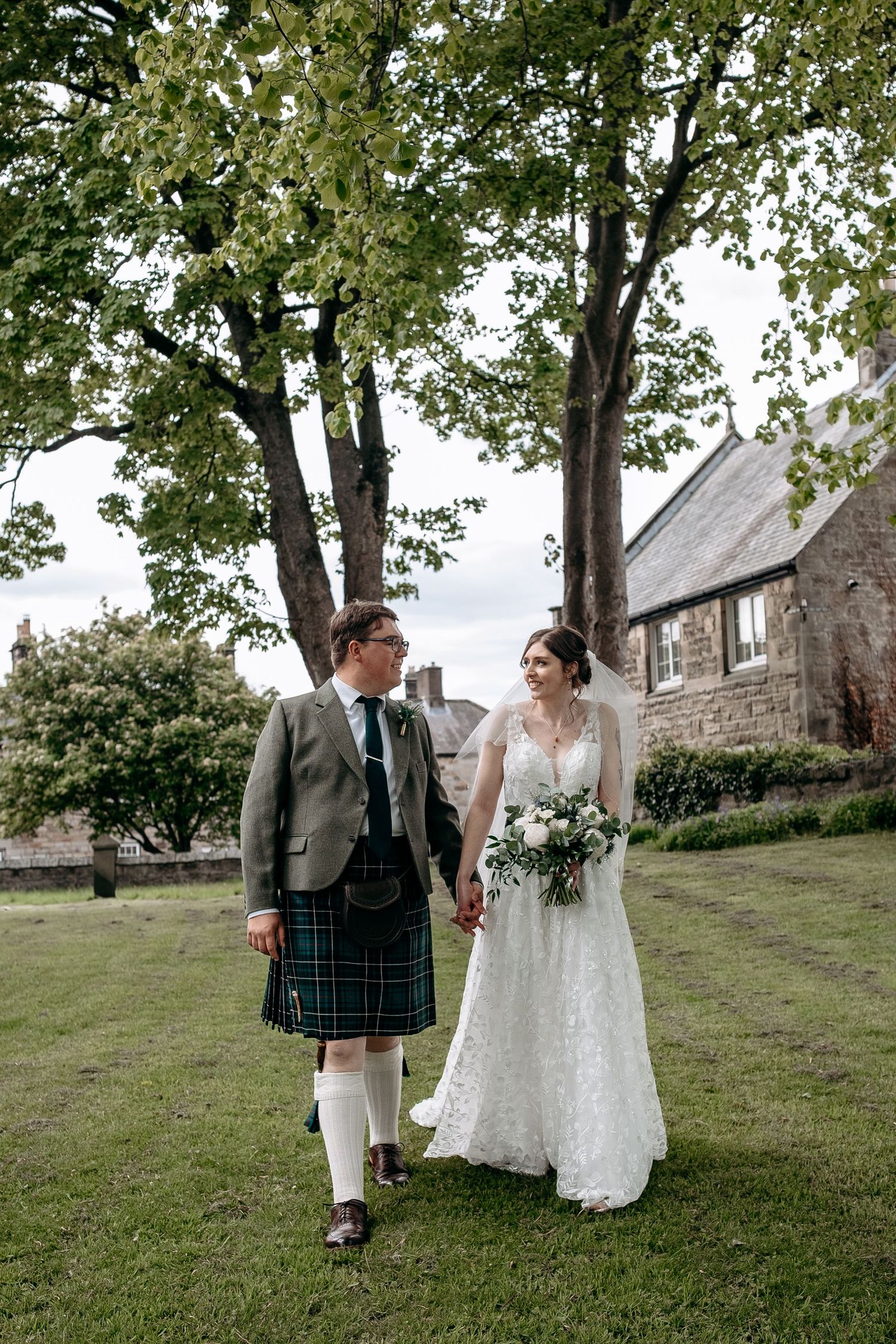 A bride and groom walk hand-in-hand on grass, groom in tartan kilt, bride in lace gown holding bouquet, stone building behind.