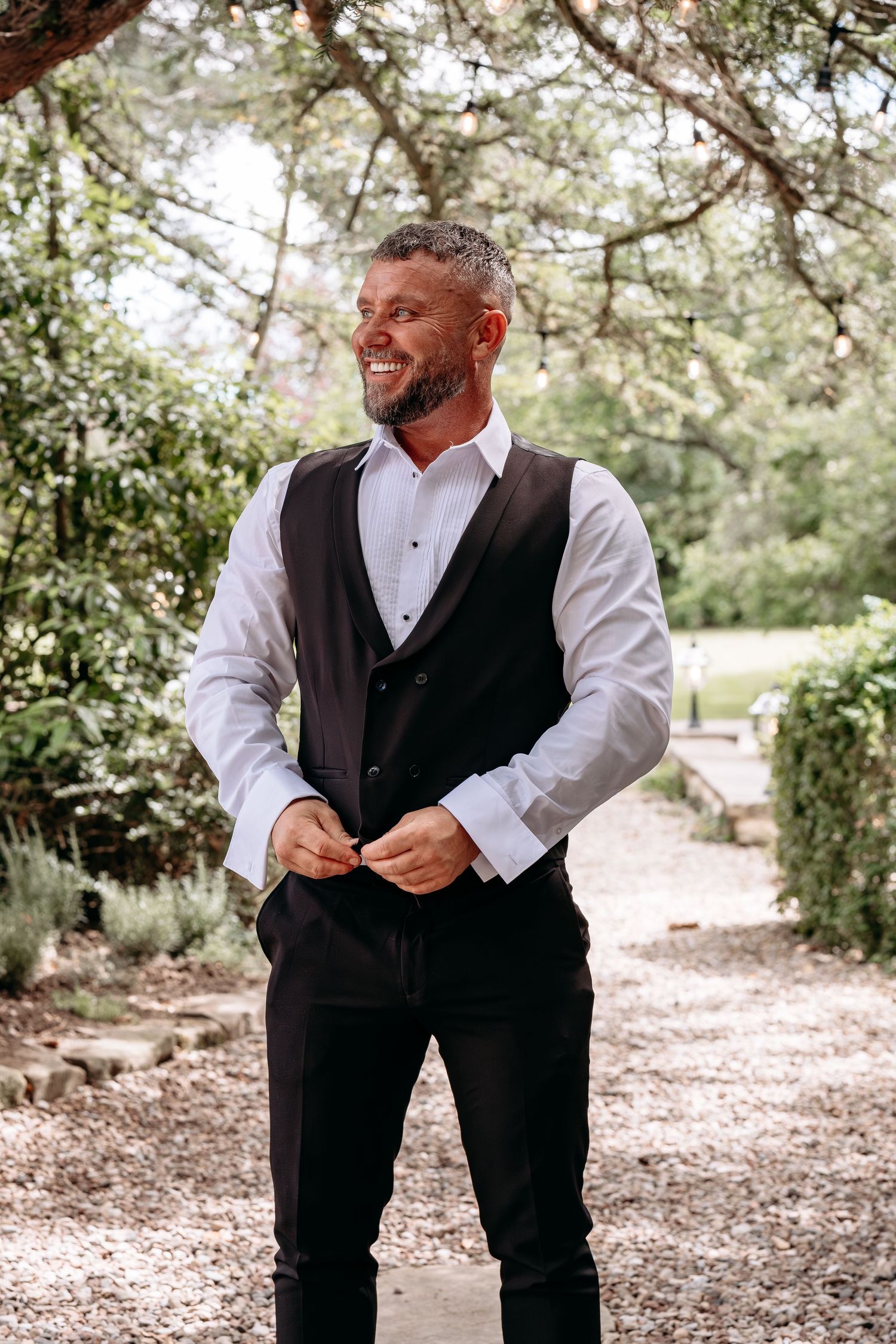 Smiling bearded groom in white shirt and black vest buttoning up outdoors in a garden setting.