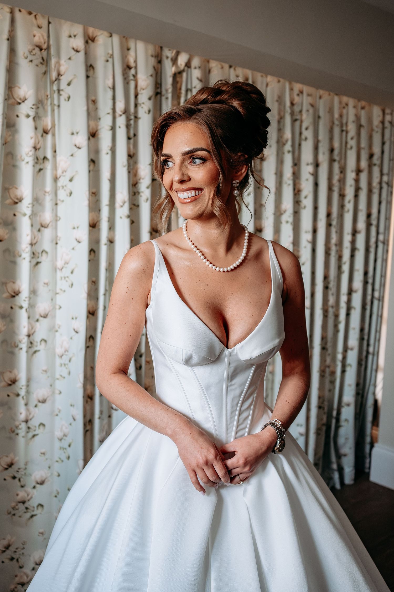 Smiling bride in white satin ballgown with pearl necklace and updo hairstyle, posing in front of floral curtains.