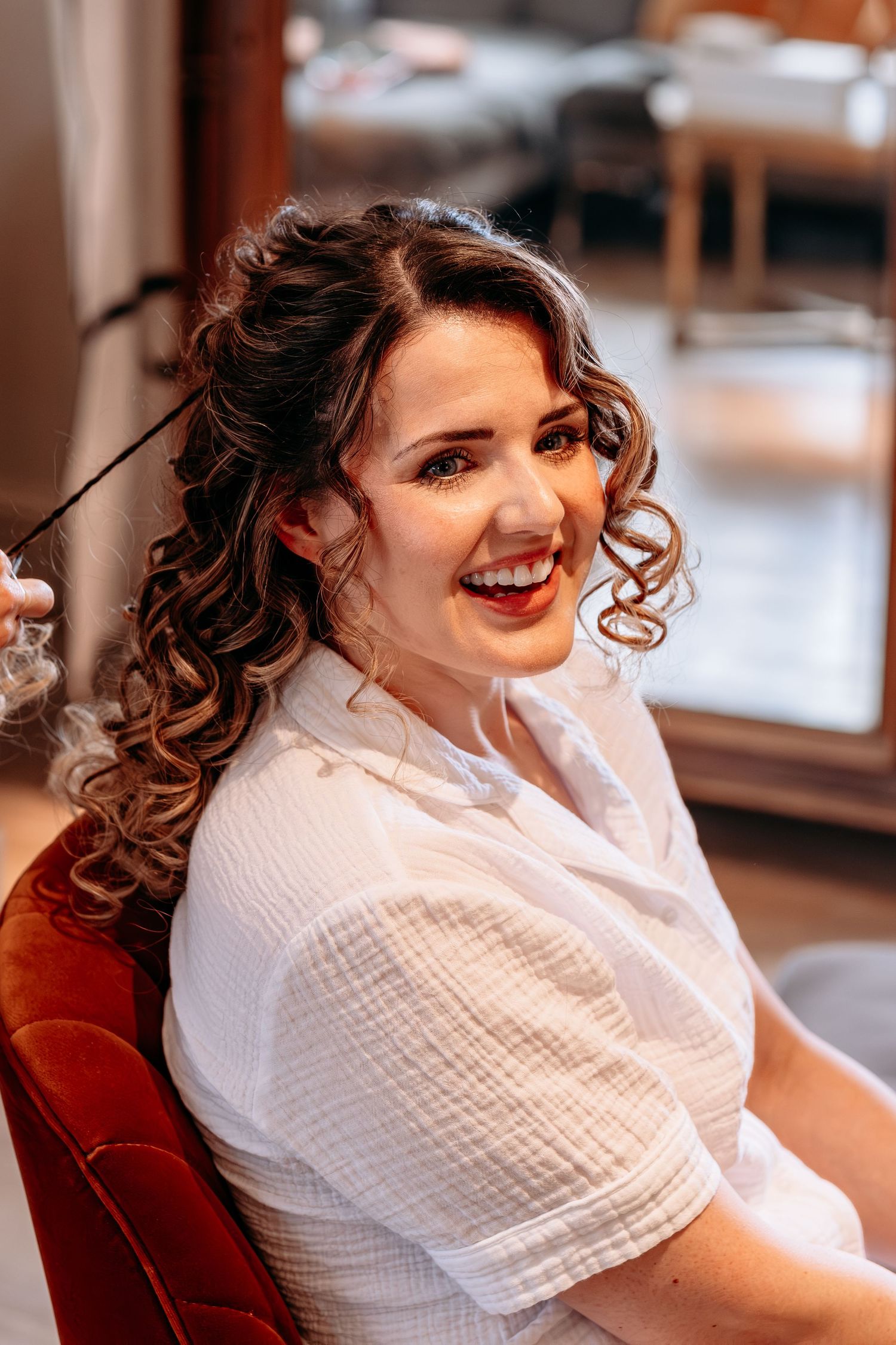 Smiling woman with curly brown hair and red lips in white top sitting in salon chair getting hair styled.