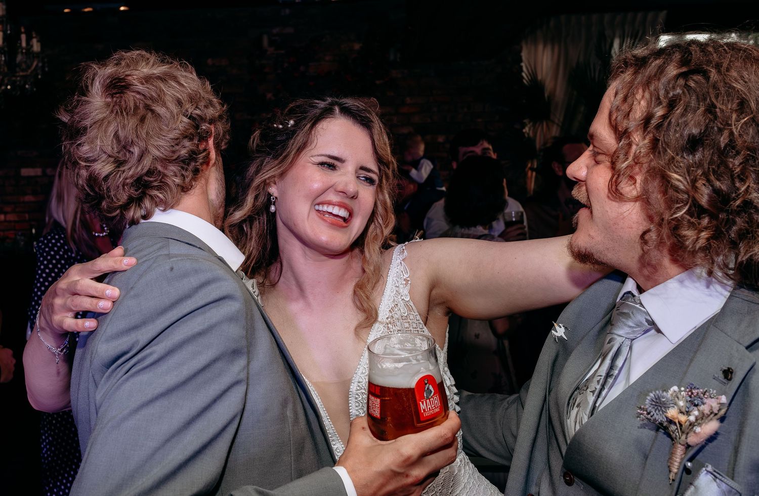 Bride in white dress laughing with guests holding a red drink at a wedding reception celebration.