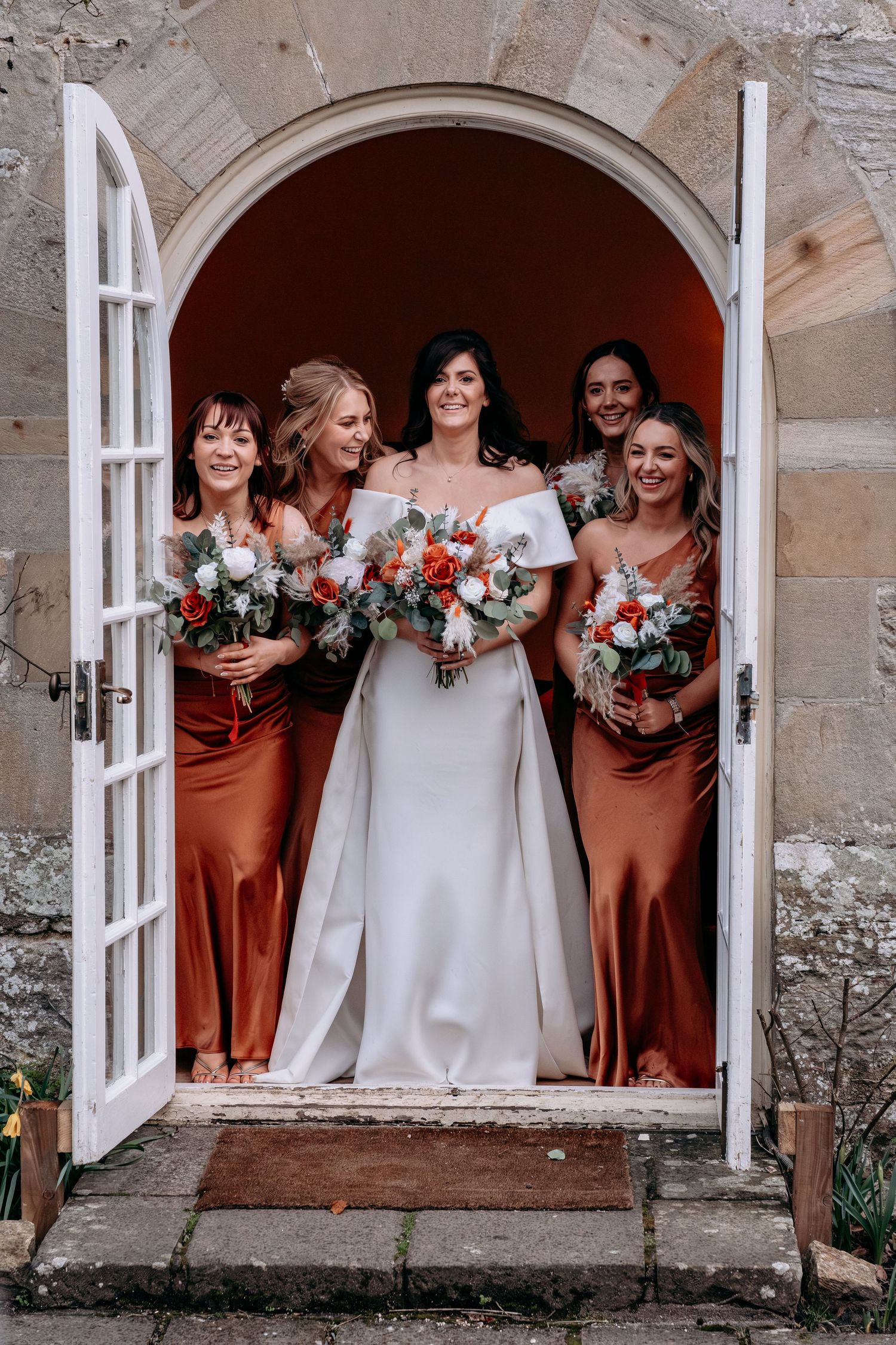 Bride in white off-shoulder gown with four bridesmaids in rust satin dresses holding bouquets in stone archway doorway.