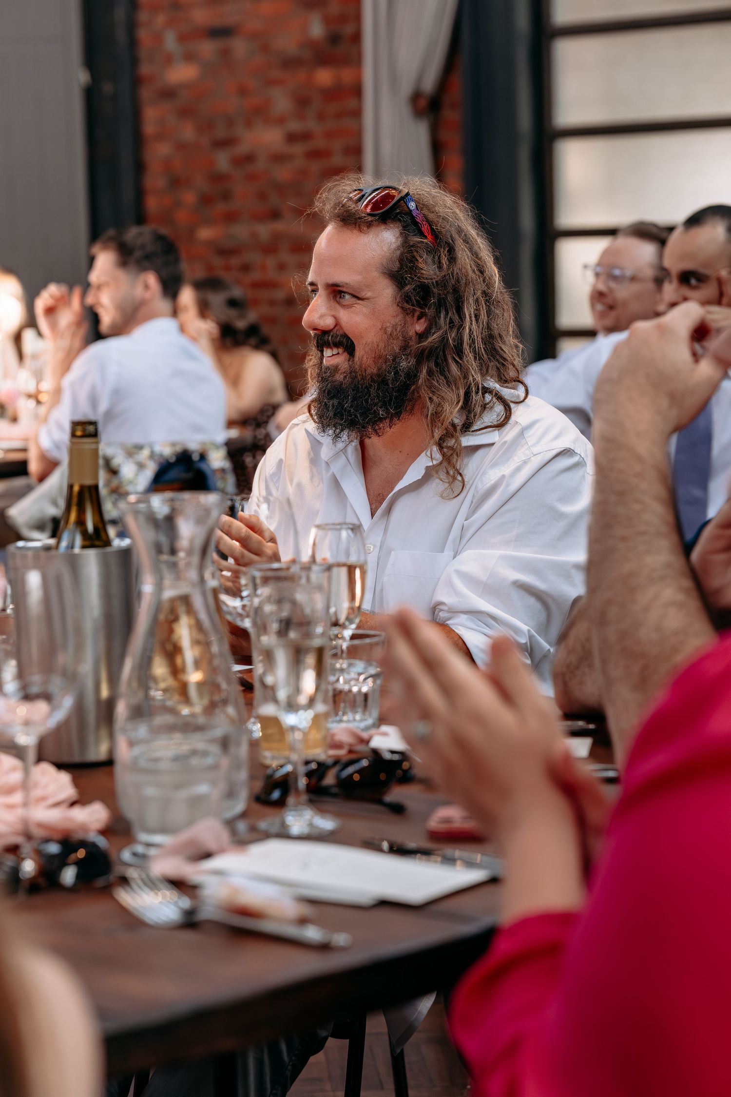 Bearded man with long curly hair smiling at a dinner party table with champagne glasses and wine bottles.
