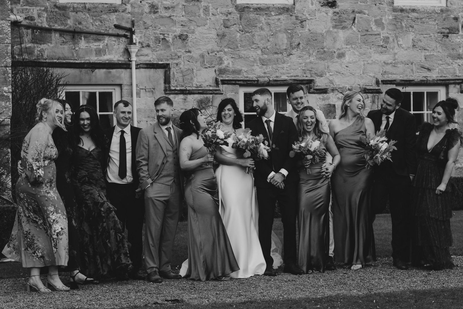 Black and white wedding party photo with bride, groom, and guests posing outside a stone building.