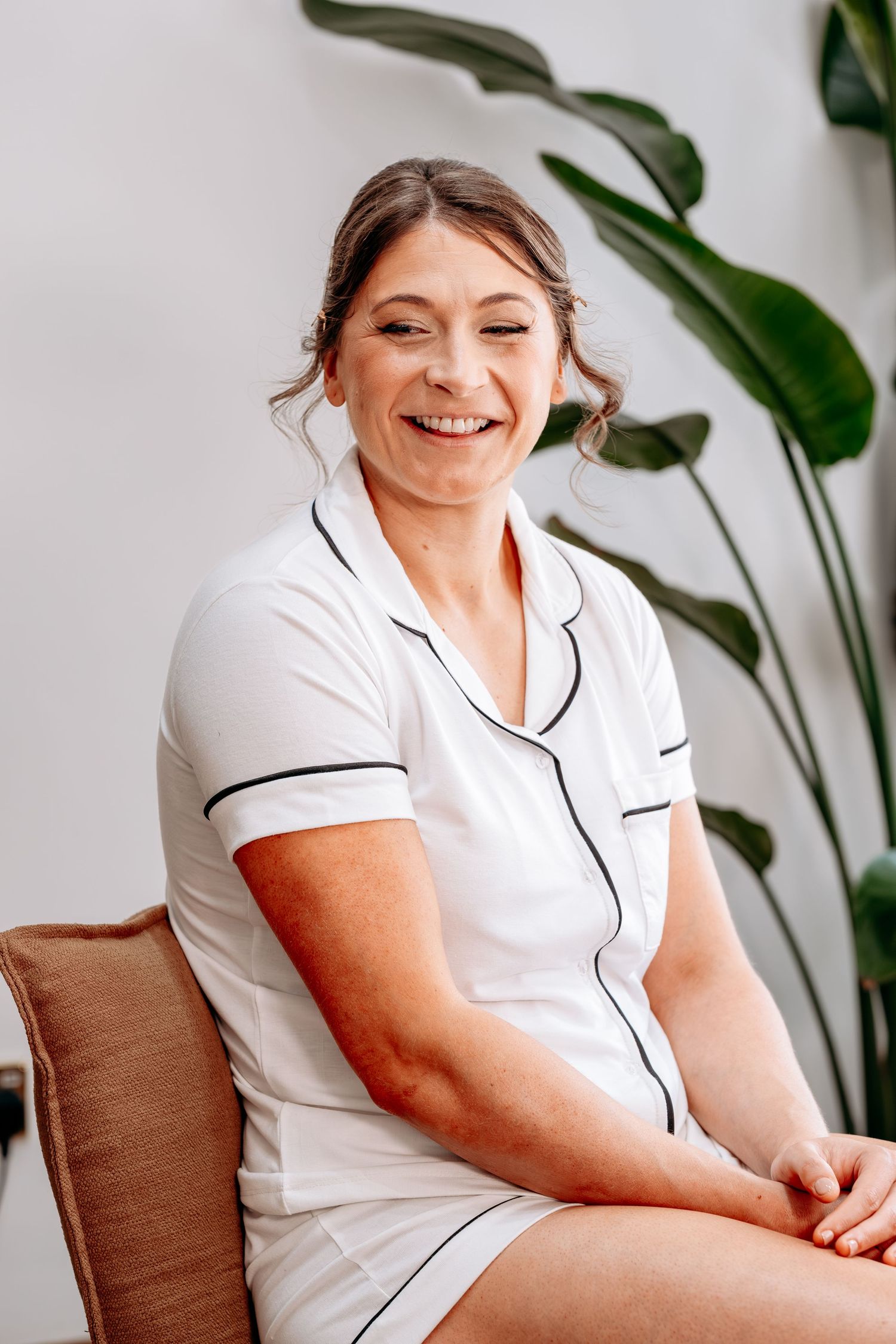 Smiling woman in white nurse uniform seated in chair with tropical plant in bright modern spa setting.