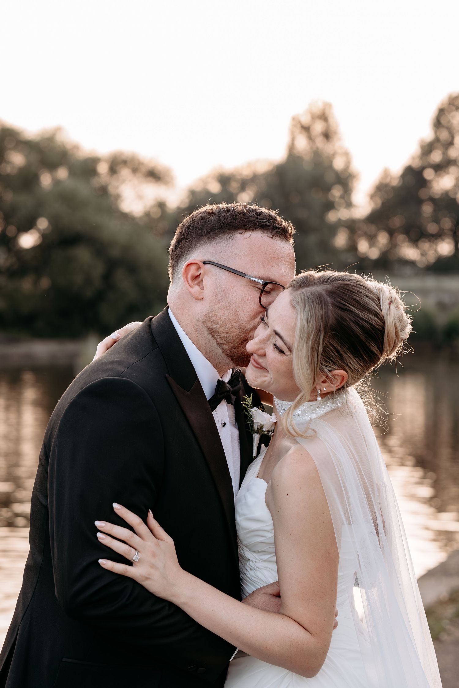 Groom in black tuxedo kisses bride on forehead near a lake at golden hour, bride wearing white dress and veil.