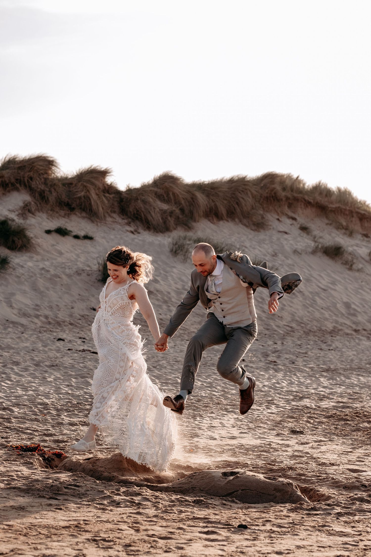 Joyful bride and groom holding hands, leaping on a sandy beach with dunes, celebrating their wedding day.