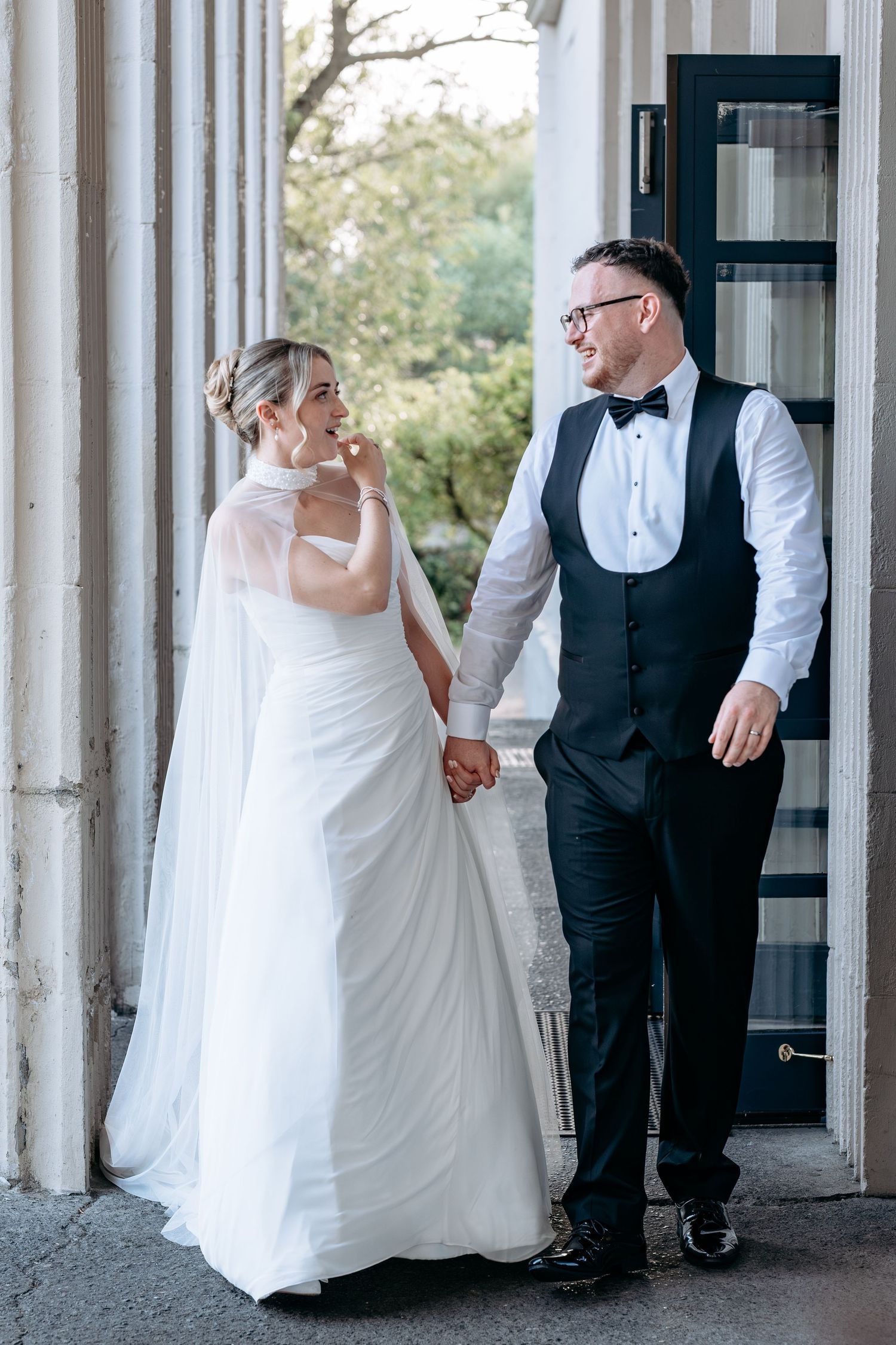 Bride in white gown and groom in black tuxedo hold hands, smiling at each other between elegant columns.